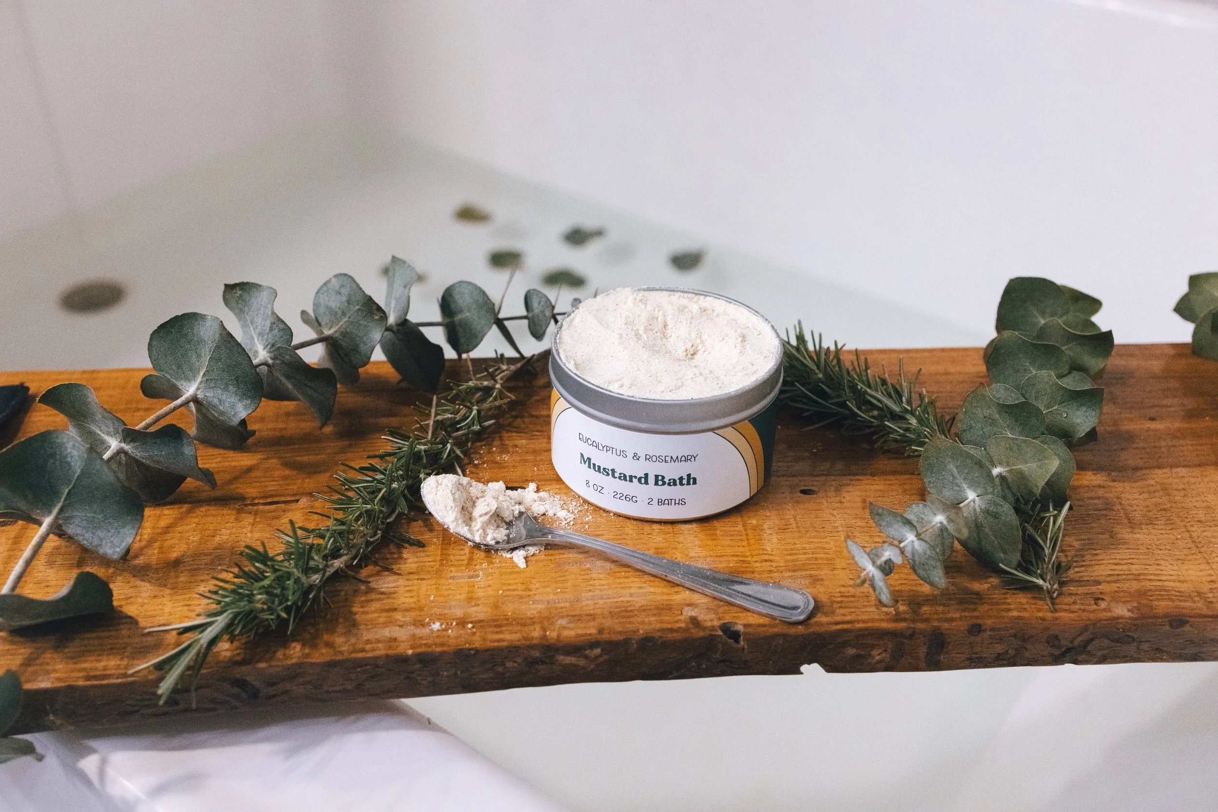 A container of mustard bath powder with a spoon on a wooden surface, decorated with eucalyptus and rosemary sprigs.