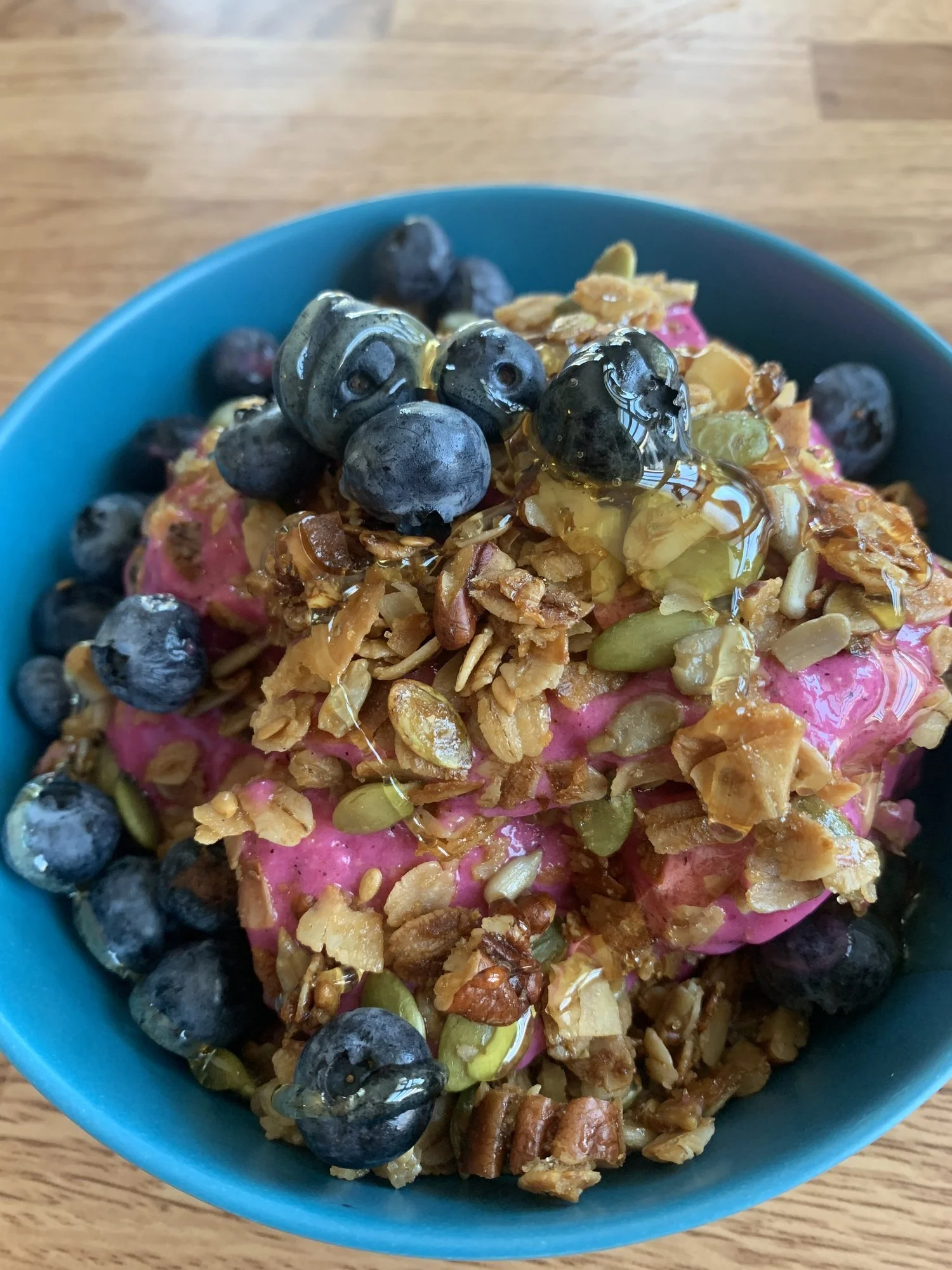 A bowl of pink frozen yogurt topped with blueberries, granola, pumpkin seeds, and honey.