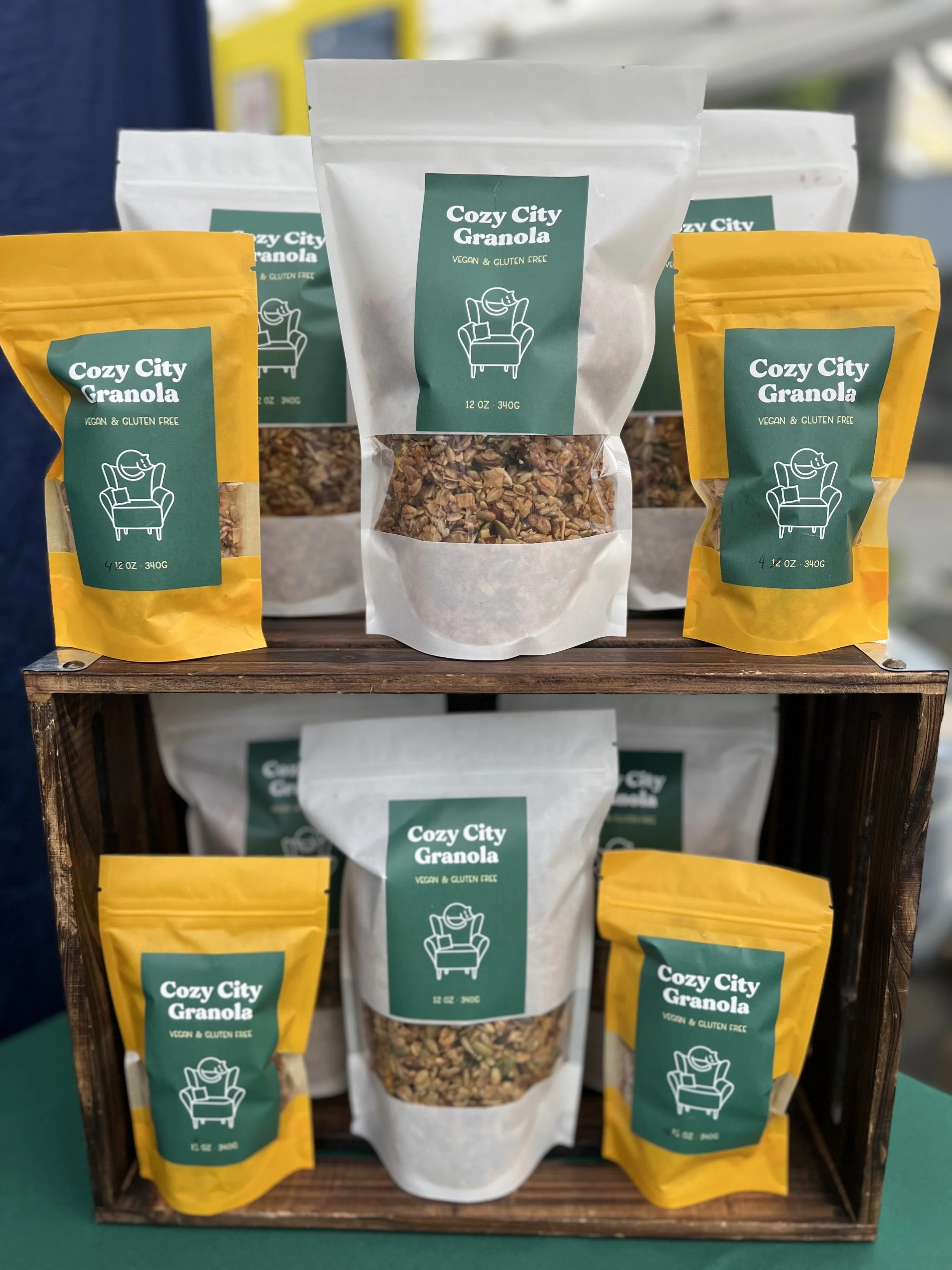Display of Cozy City Granola packages, with white and yellow bags on a wooden stand, showing label with a chair icon, cost, and product details.