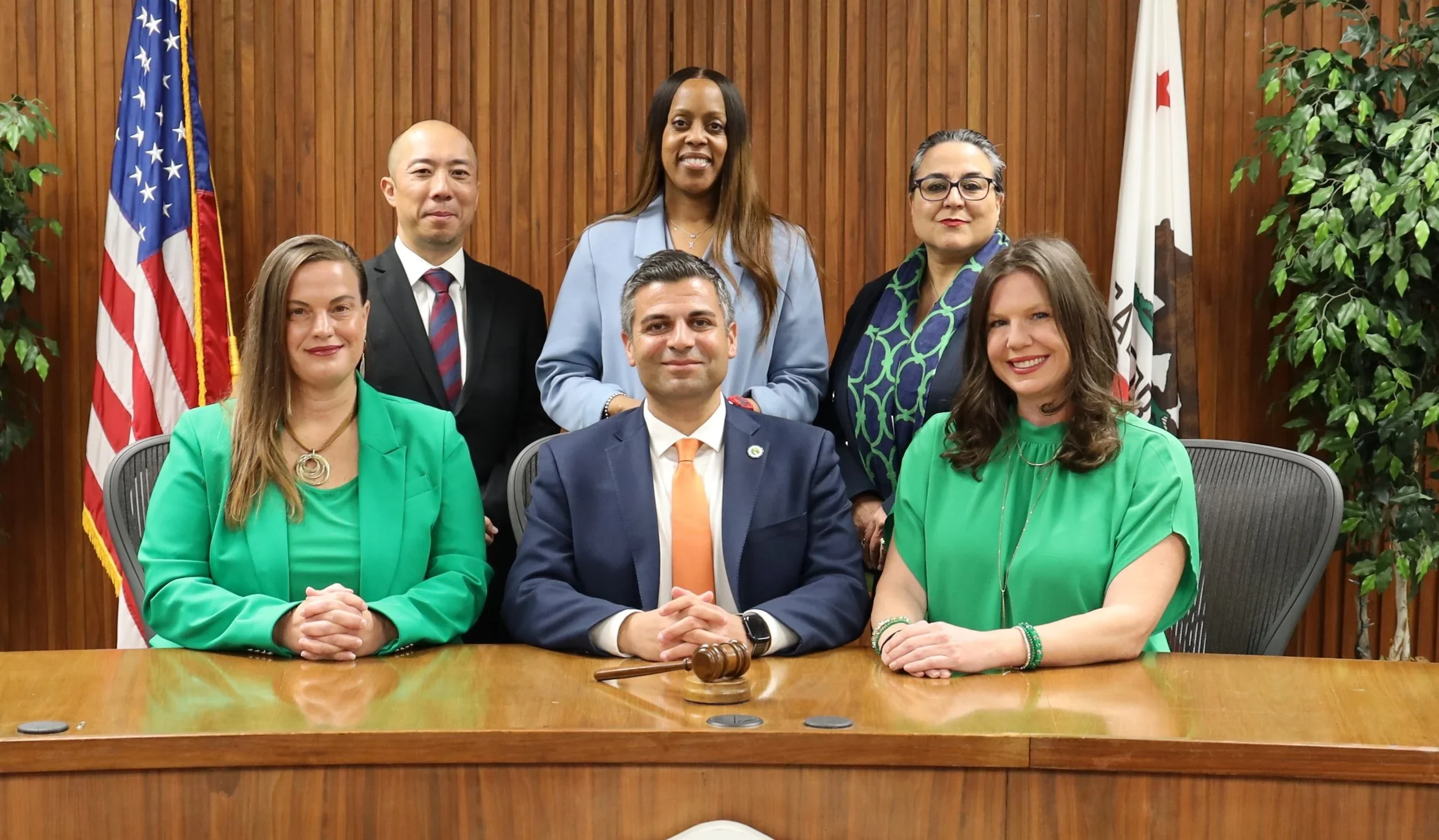 GUSD School Board portrait 2022 in a formal setting with an American flag and a California state flag behind them. They are seated and standing around a wooden conference table, dressed in business attire, smiling for the photo.