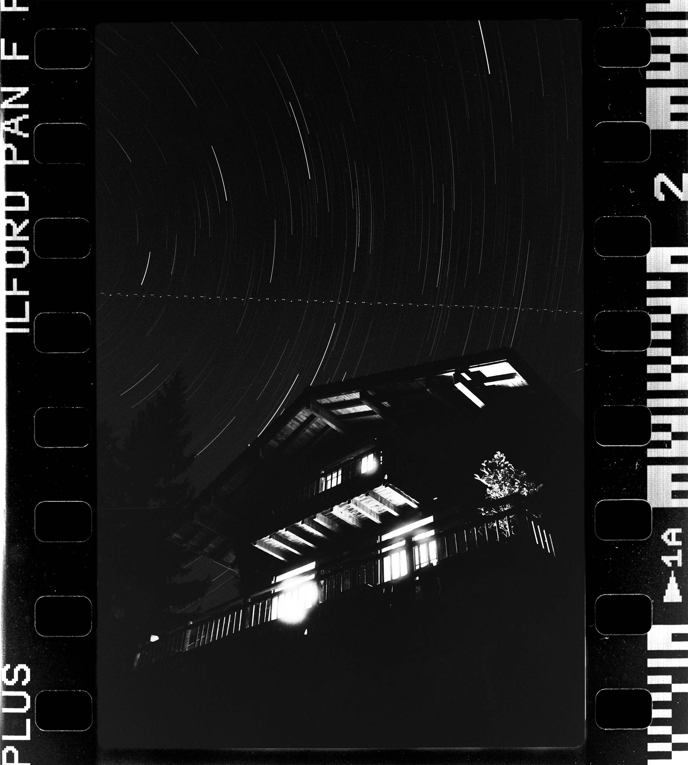 Nighttime photograph of star trails above a wooden building with a sloped roof and open door, with a tree visible in the lower right corner.