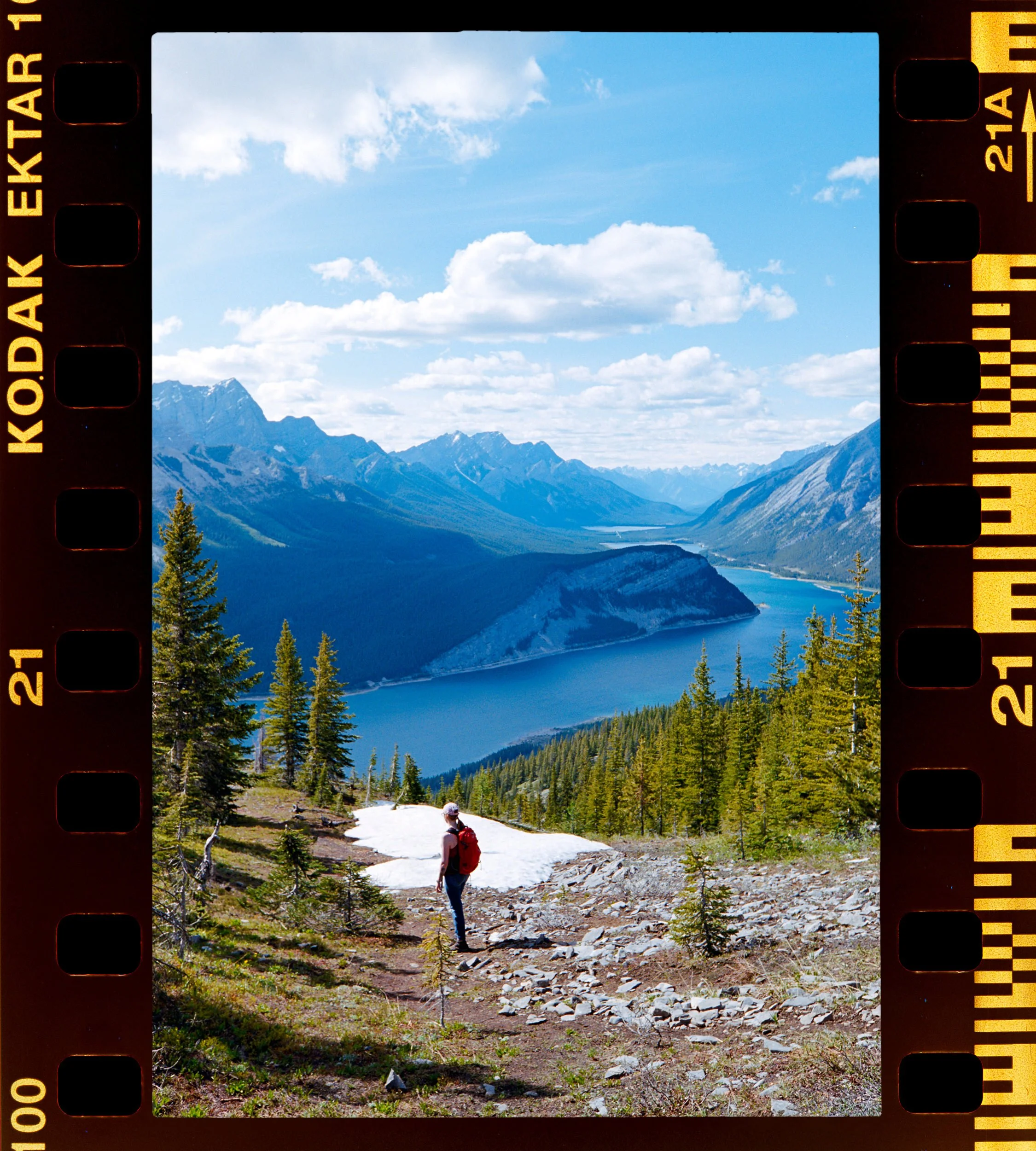 Person with a red backpack hiking on a rocky trail surrounded by green trees, with mountain peaks and a blue lake in the background under a partly cloudy sky.