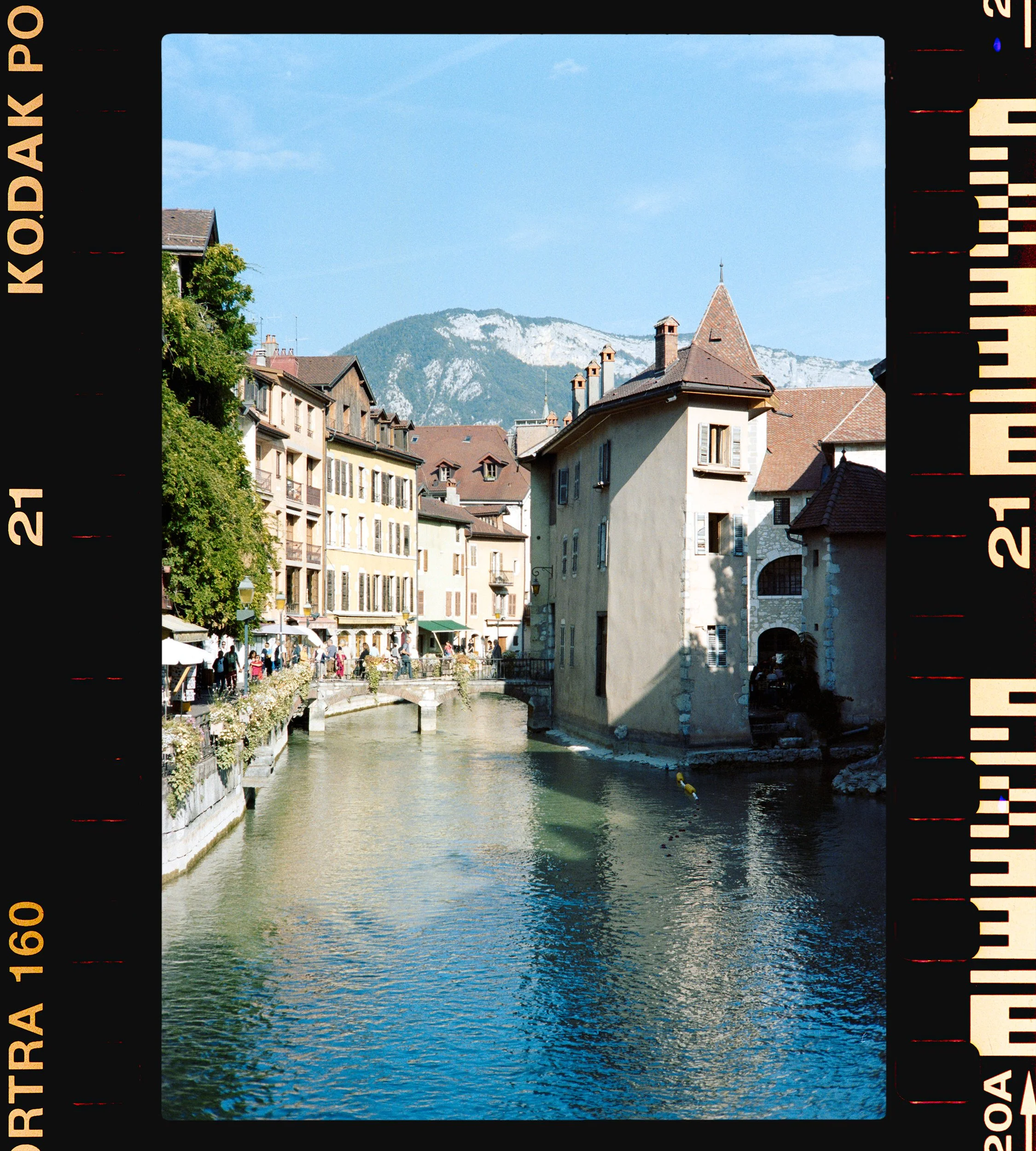 A European town with colorful buildings along a narrow river, mountains in the background, and people walking on the riverside walkway on a sunny day.