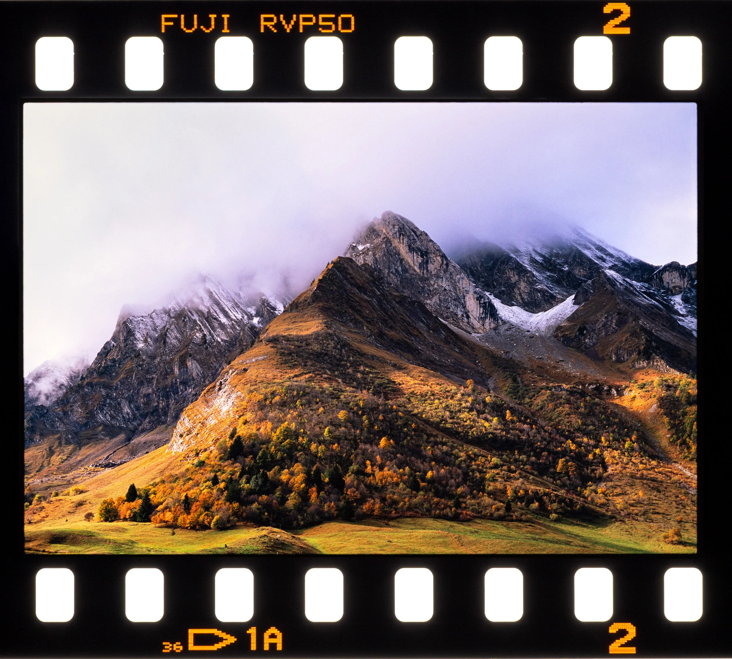 A photograph of a landscape with tall mountains, some covered in snow, surrounded by clouds and mist. The foreground shows a grassy area with patches of yellow and green trees.