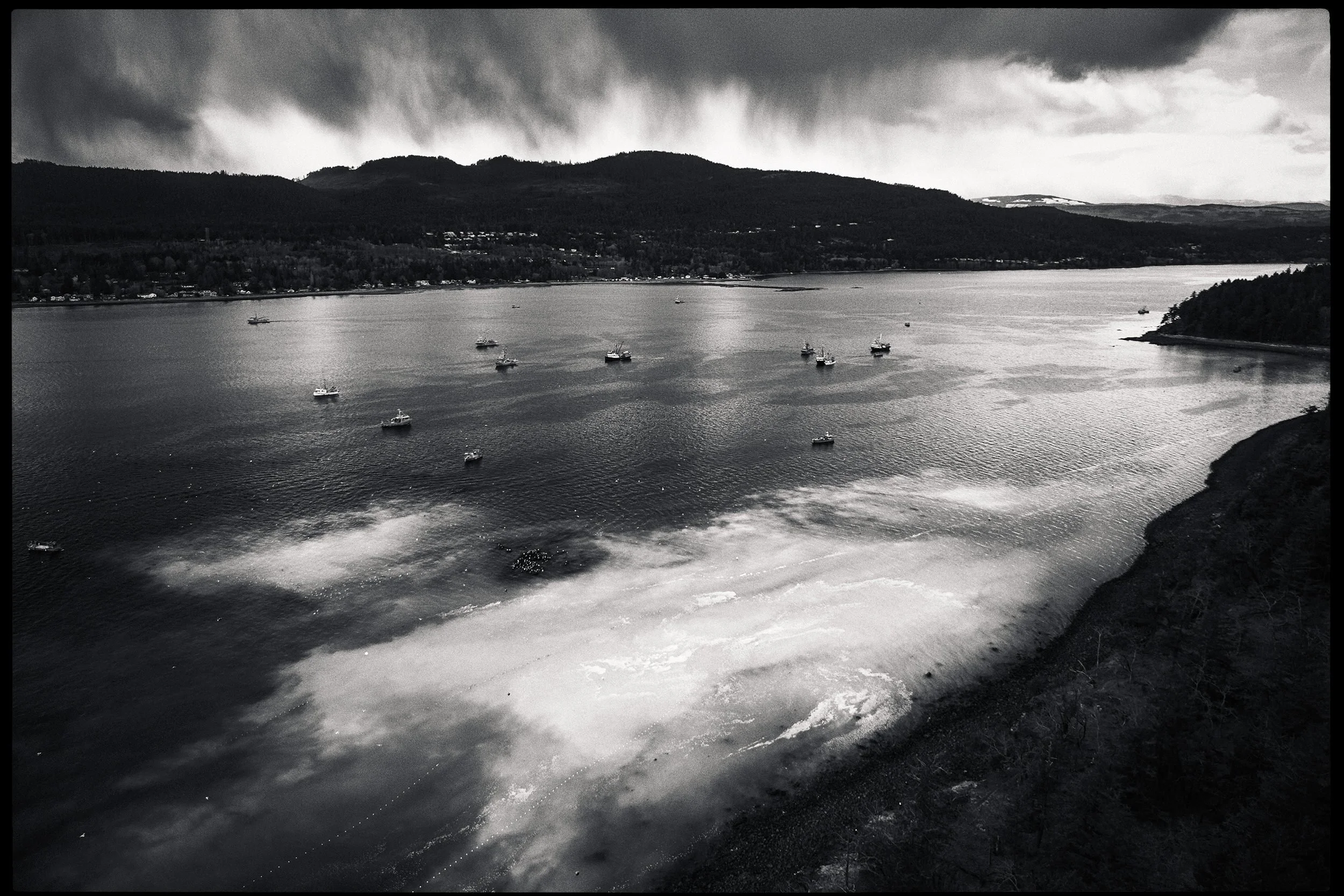 Black and white photo of a lake with boats and a mountainous shoreline in the background, under a dark, cloudy sky.