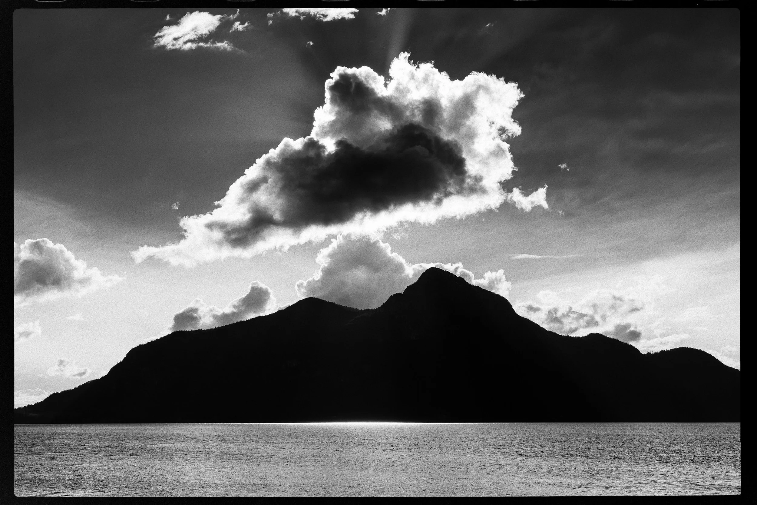 Black and white photo of a mountain range with a body of water in the foreground, large cloud formation in the sky with sunlight rays emerging from behind.