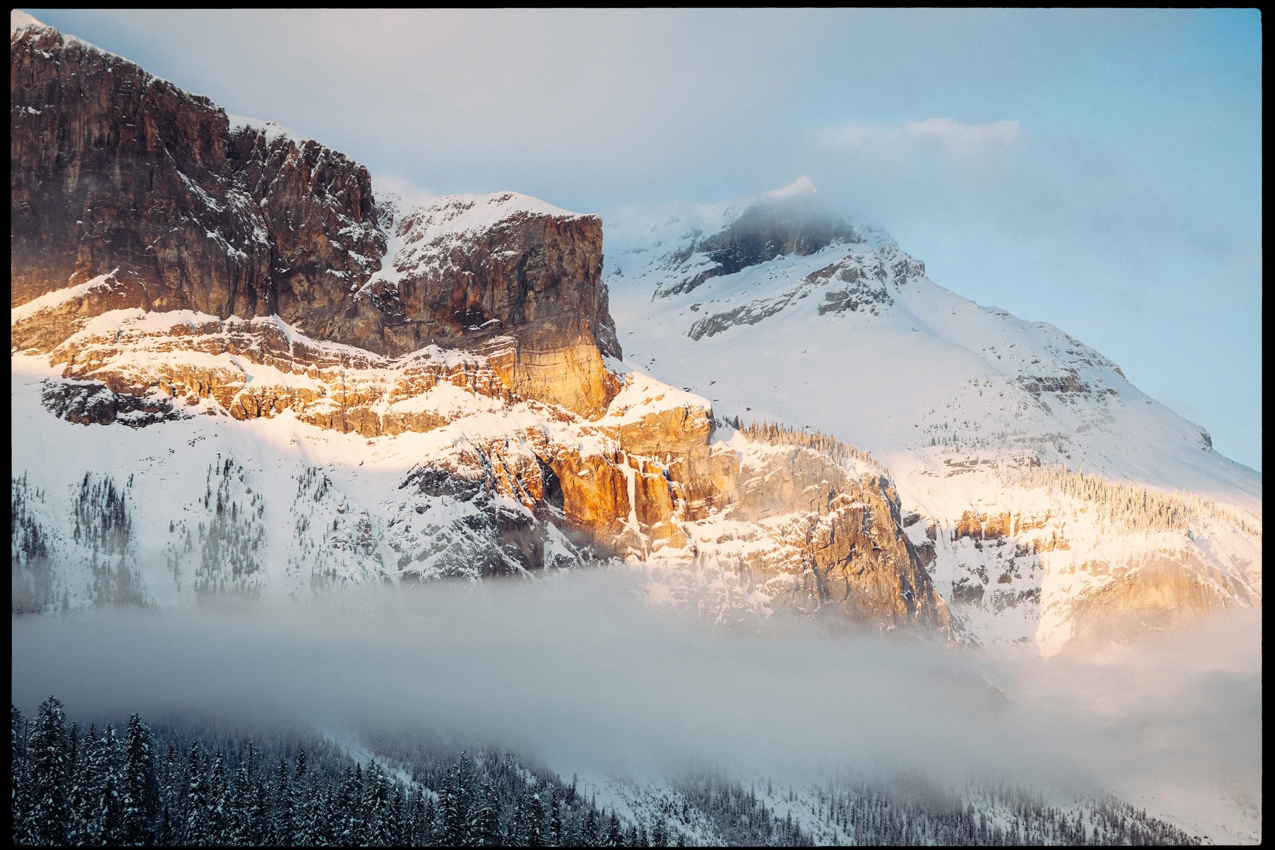 Snow-covered mountain peaks with rocky cliffs illuminated by sunlight, dense pine forest at the base, and some clouds partially covering the mountain tops.