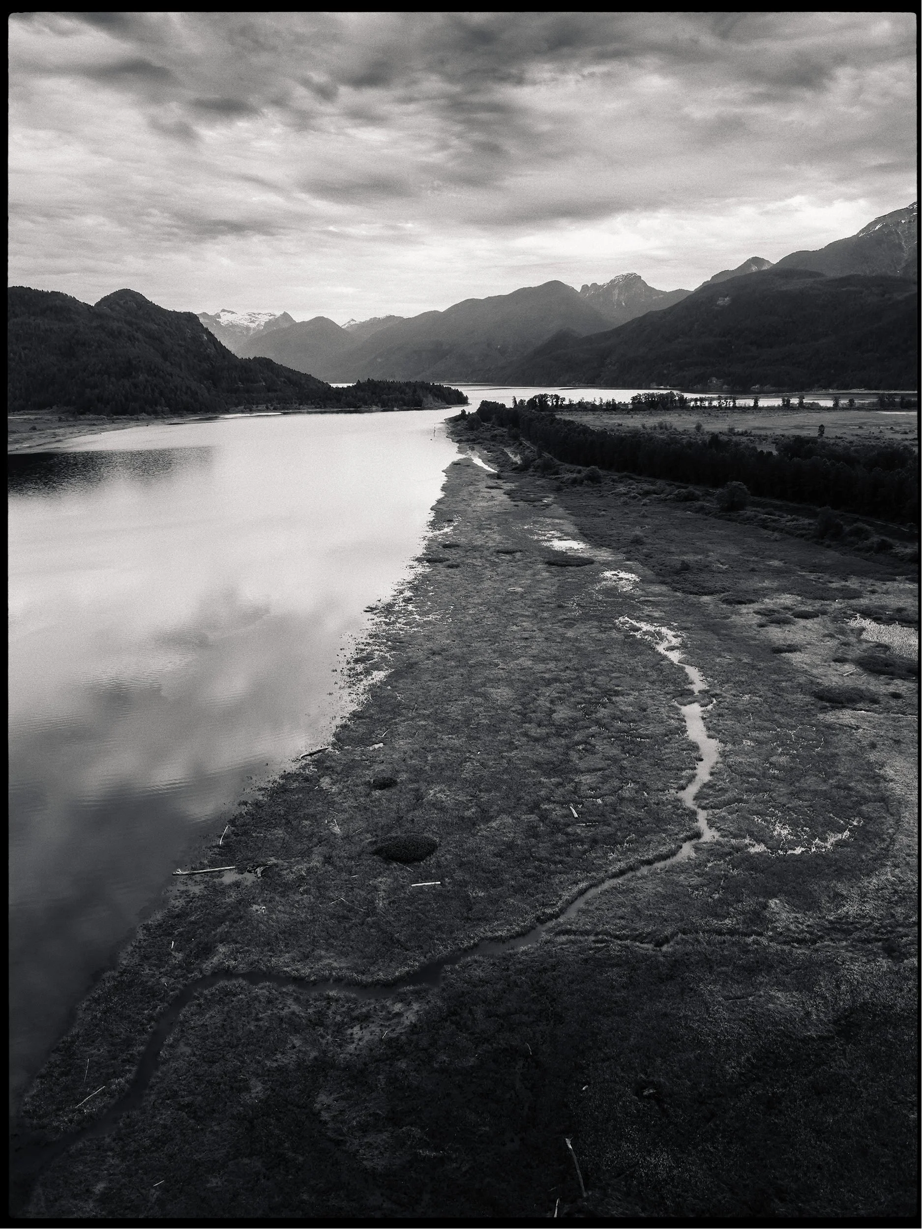 Black and white landscape photograph of a river with mountains in the background and a cloudy sky.
