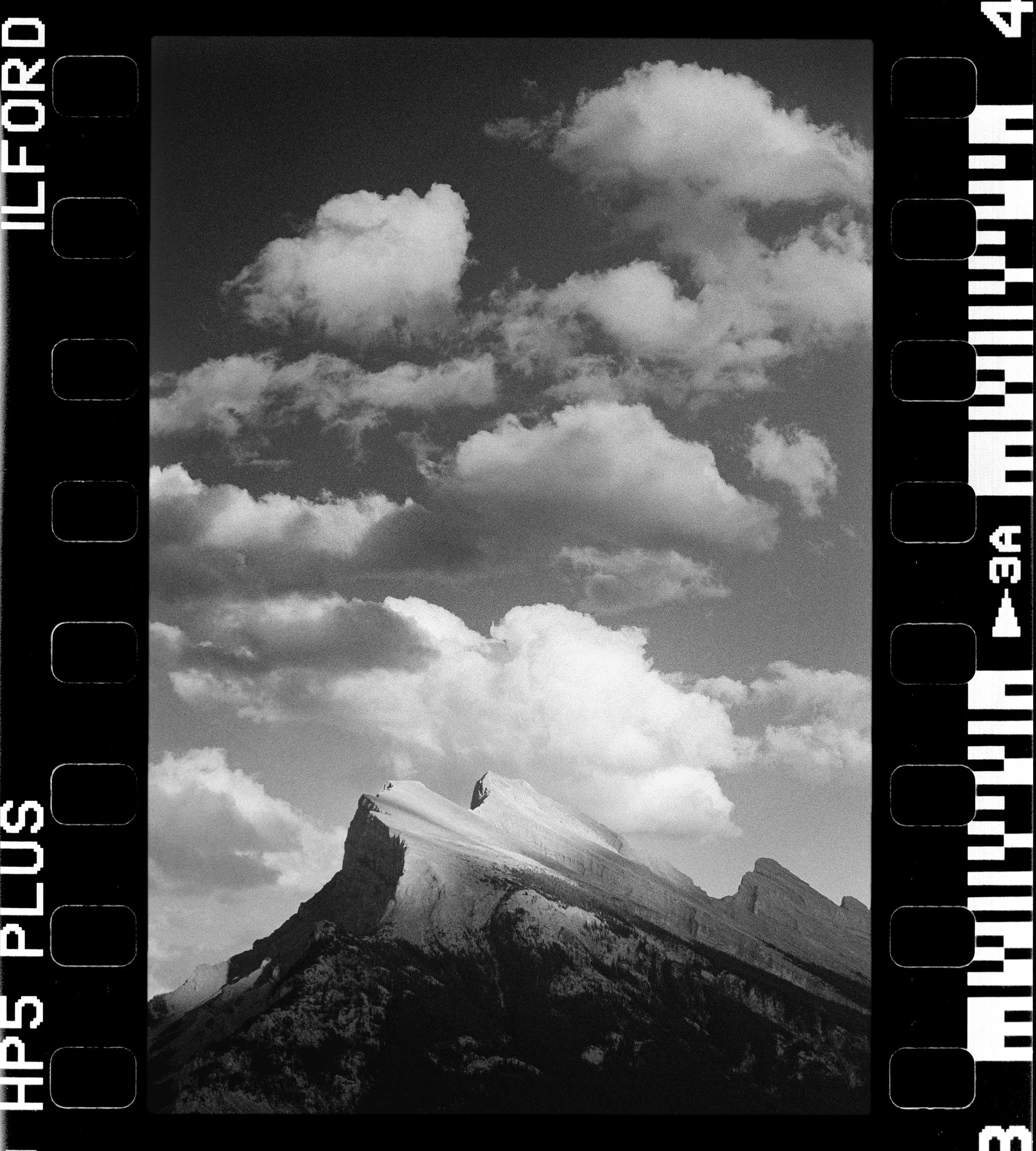 Black and white photograph of a mountain with a snow-capped peak and rugged cliffs, under a cloudy sky.