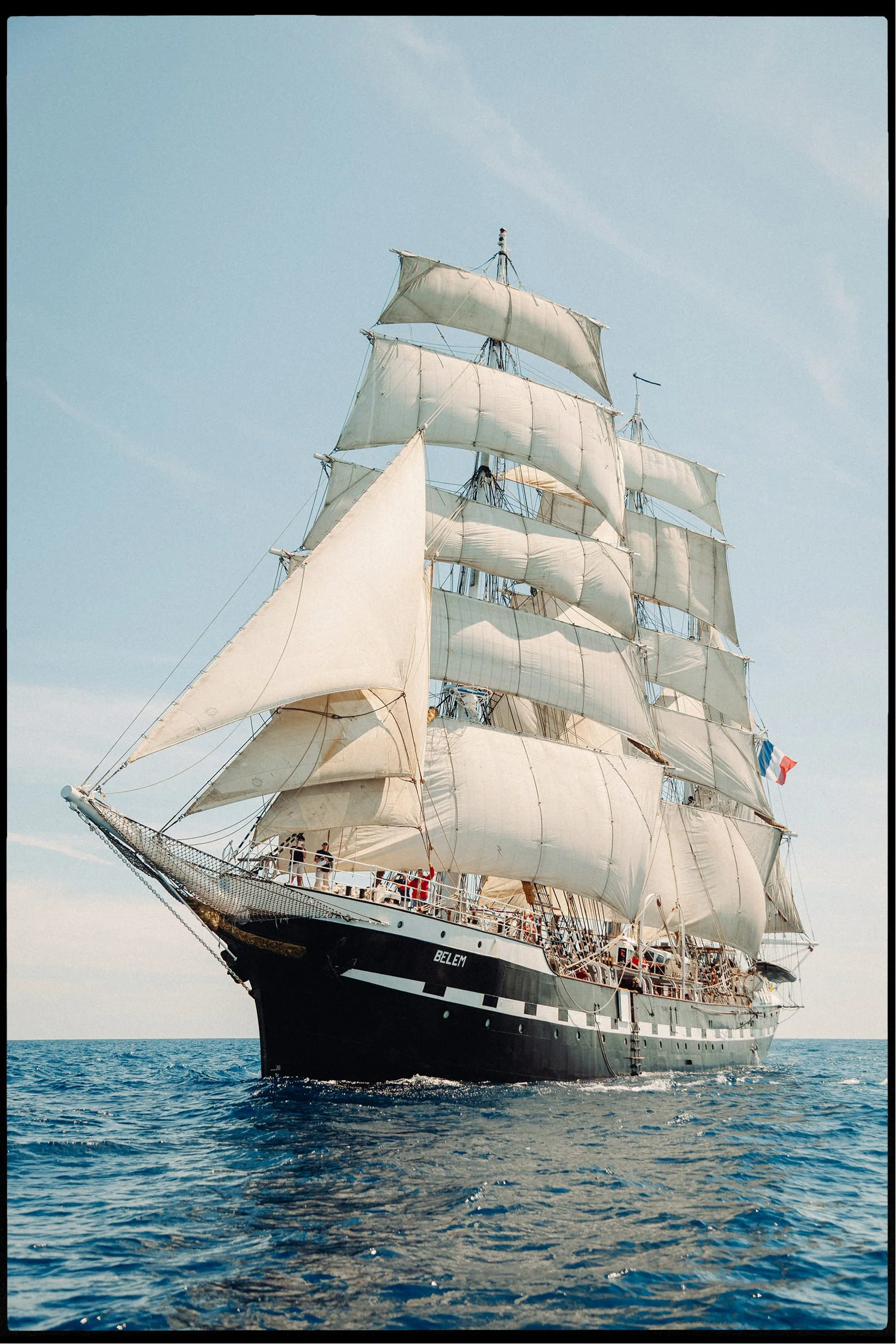 A large sailing ship with white sails and a black hull named 'BELEM' on the open water under a clear blue sky.