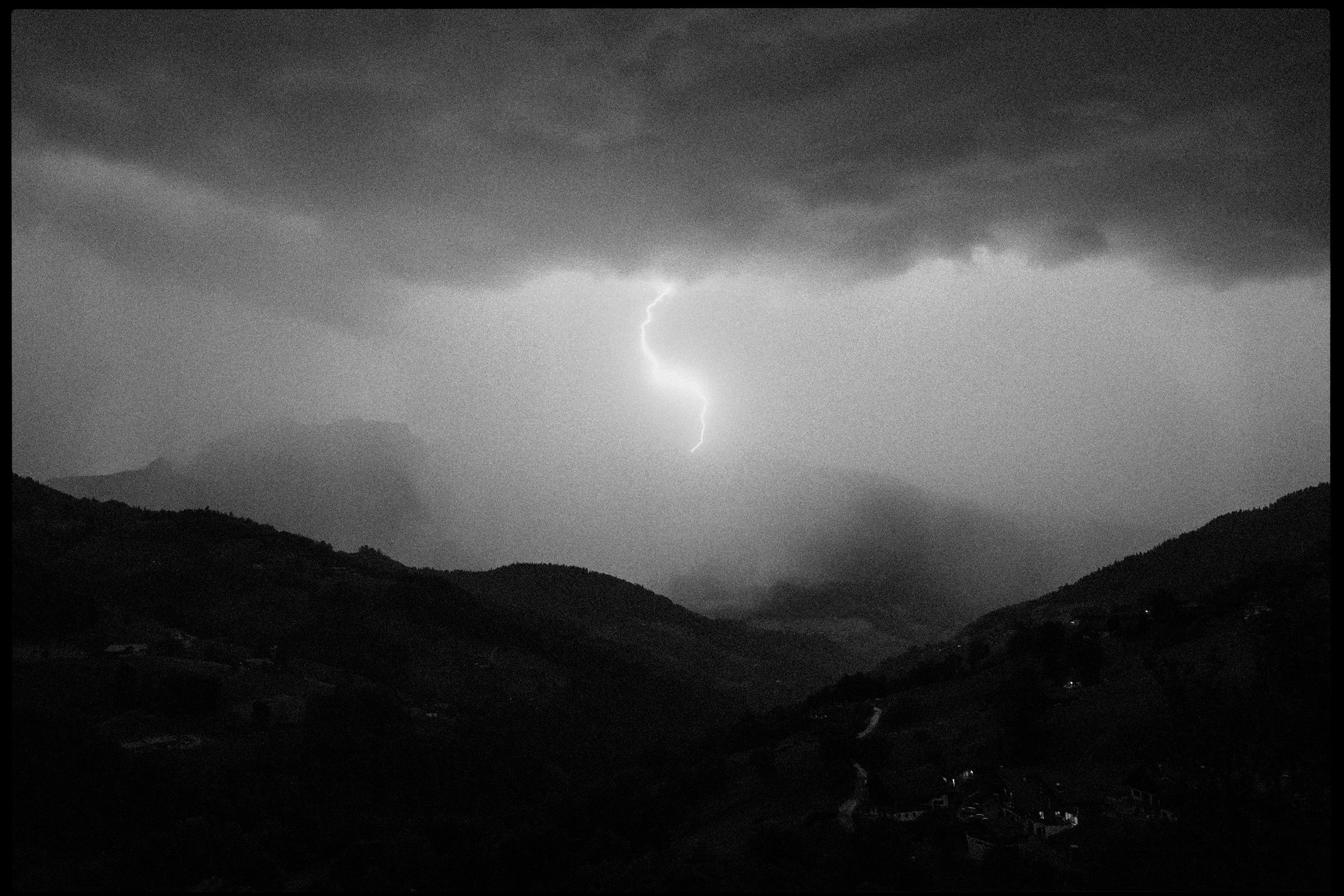 Black and white photo of a lightning bolt striking down from dark storm clouds over hilly landscape.