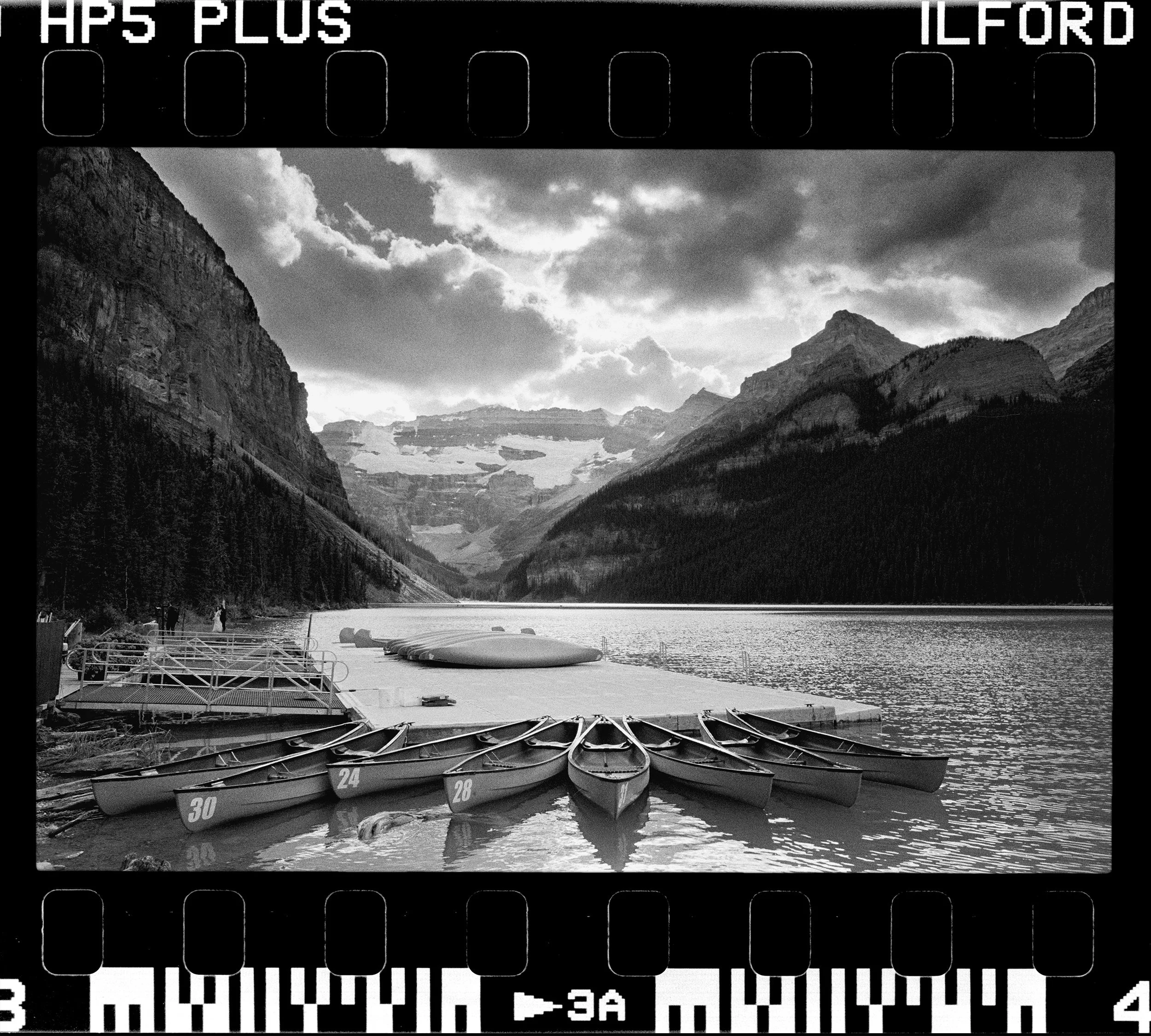 Black and white photo of boats docked along a lake, with mountain scenery and cloudy sky in the background. There are numbered boats and paddles on the shore.