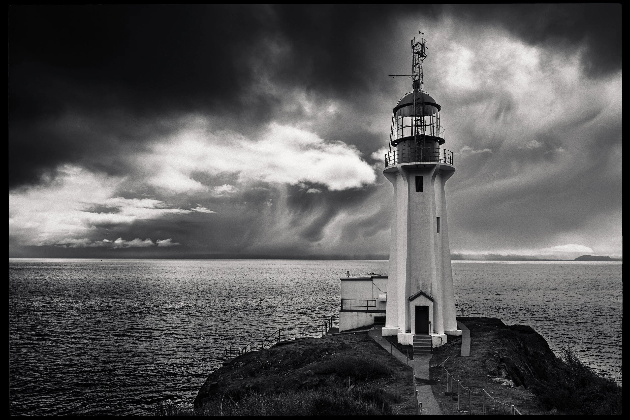 A black and white photograph of a lighthouse situated on a rocky coastline, with ocean and a distant landmass in the background under a cloudy sky.