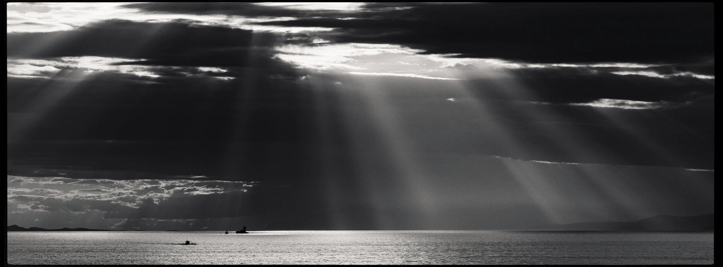 Sun rays breaking through dark clouds over the ocean, with small boats and distant land on the horizon in black and white.