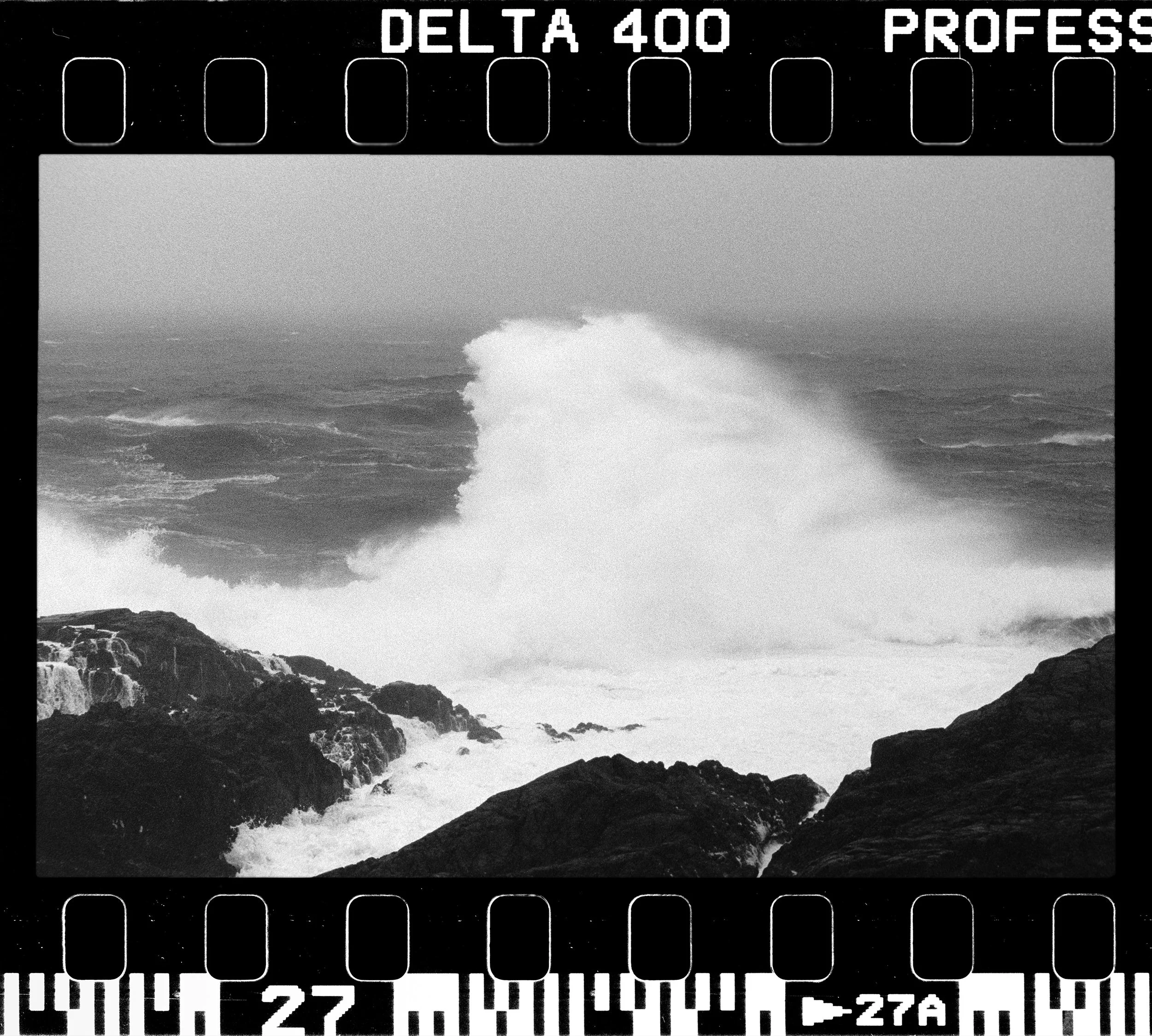 Black and white photograph of ocean waves crashing against rocky shoreline with sea spray, overlaid with film strip graphics, some text reading 'DELTA 400 PROFESSIONAL' at the top.