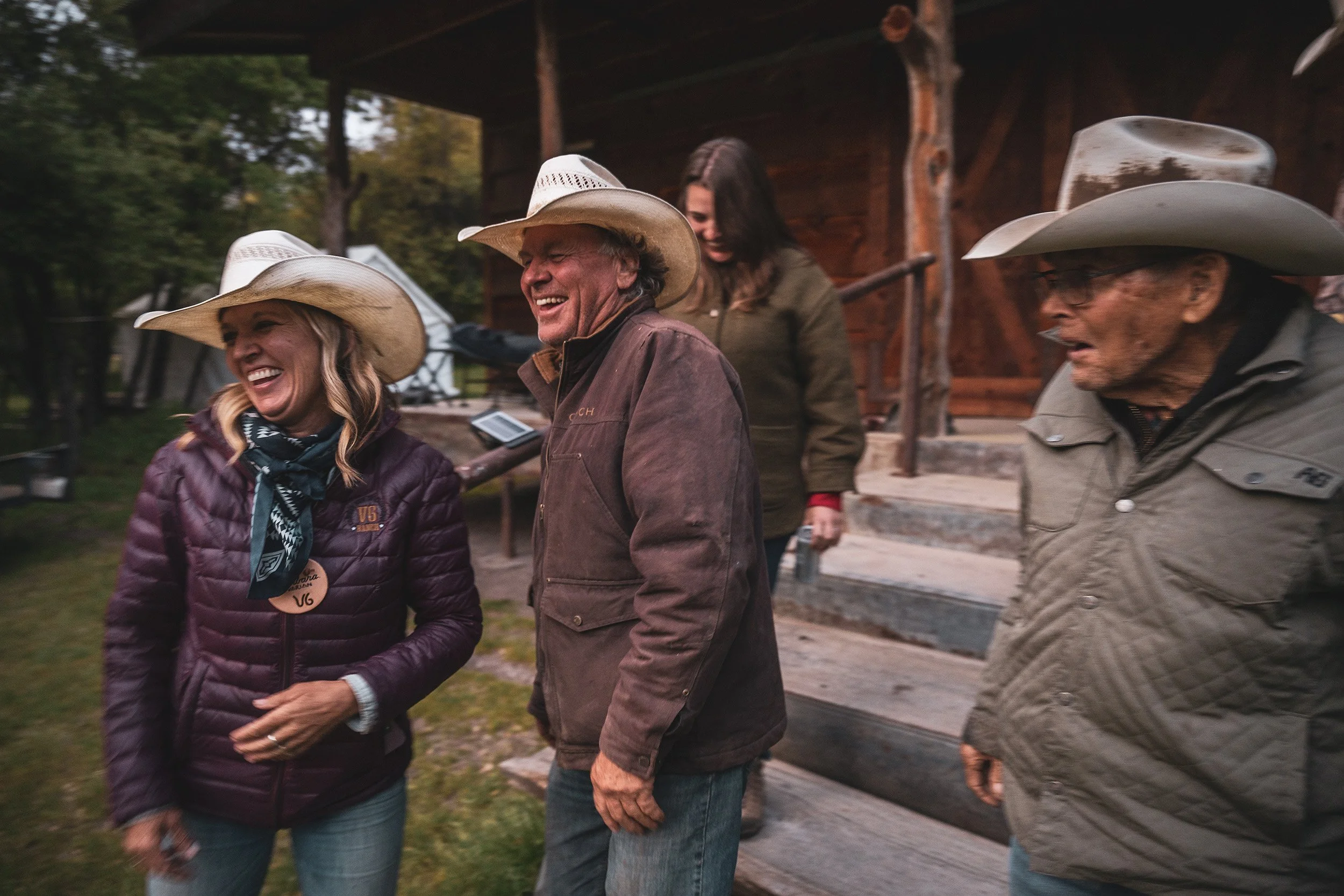 Group of four adults wearing cowboy hats, smiling and laughing outdoors near a wooden building, with trees and tents in the background.