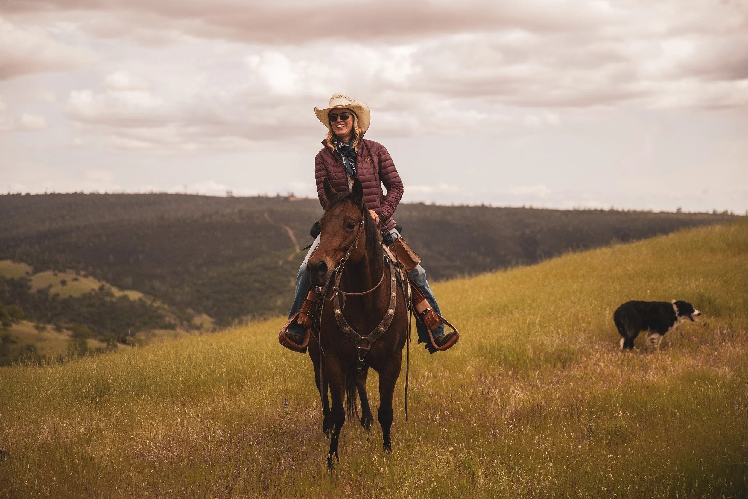 A woman riding a horse in a grassy field with rolling hills and cloudy skies in the background, wearing a wide-brimmed hat and sunglasses, and smiling.