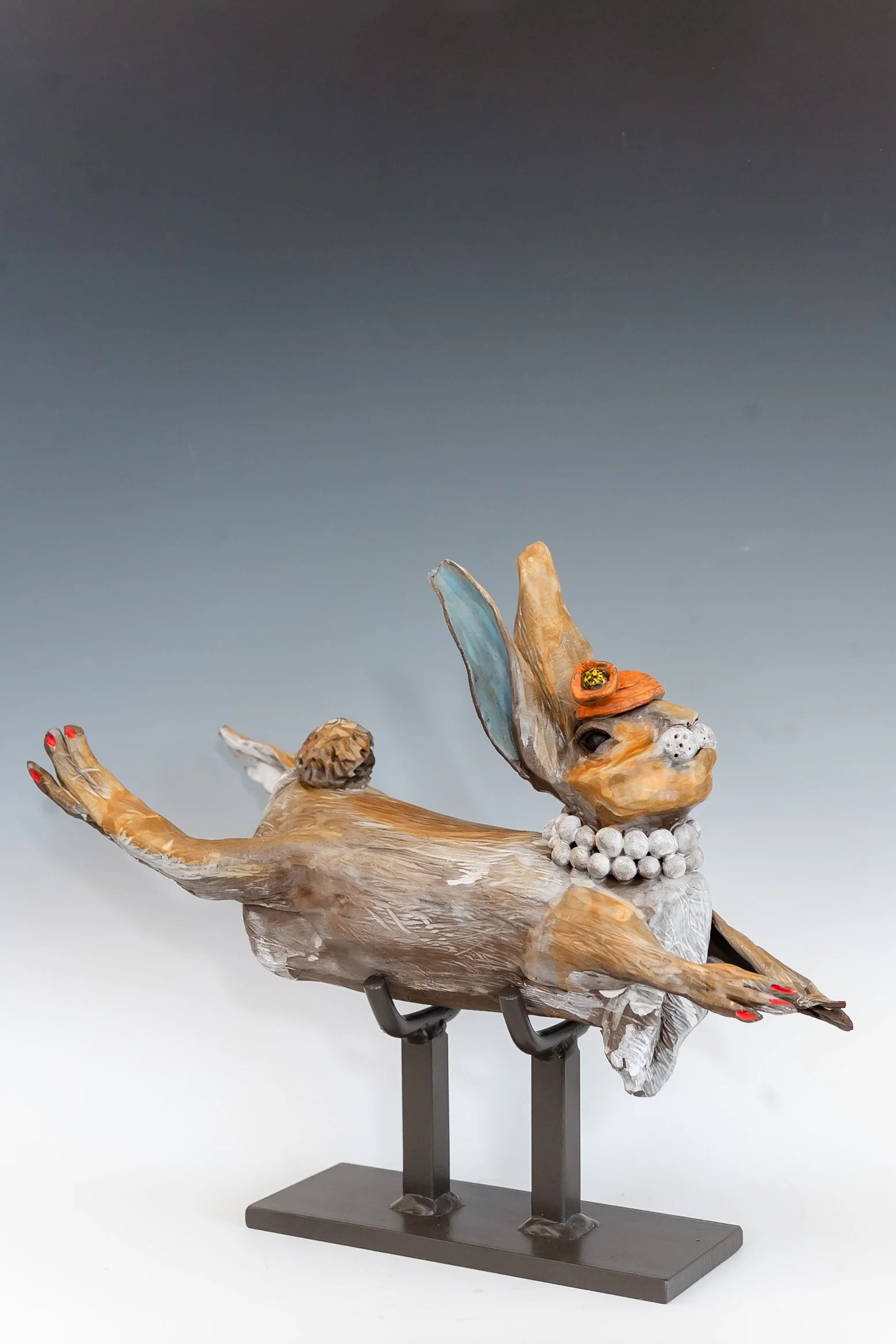 A whimsical sculpture of a rabbit with one ear upright and the other drooping, wearing a pearl necklace and an orange hat, with red painted nails, displayed on a black stand against a gradient background.