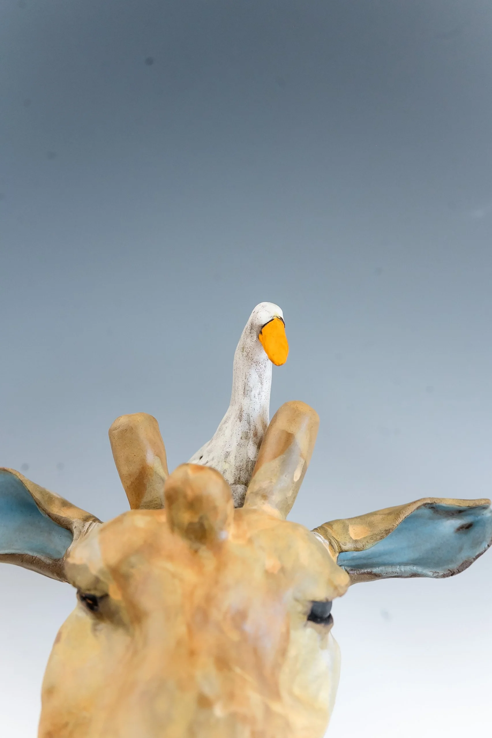 Close-up of a ceramic sculpture of a giraffe with a white bird perched on its head, against a plain background.