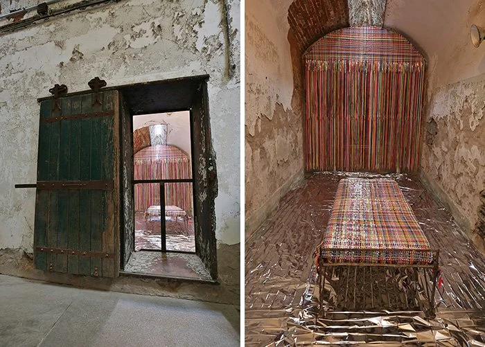 Deadly Weapons (outside & inside views), Eastern State Penitentiary installation, 2021-25, Steel frame and cot, mylar sheets, shoelaces and mylar strips for woven blanket and wall hanging, cell dimensions: 216” x 96” x 108”