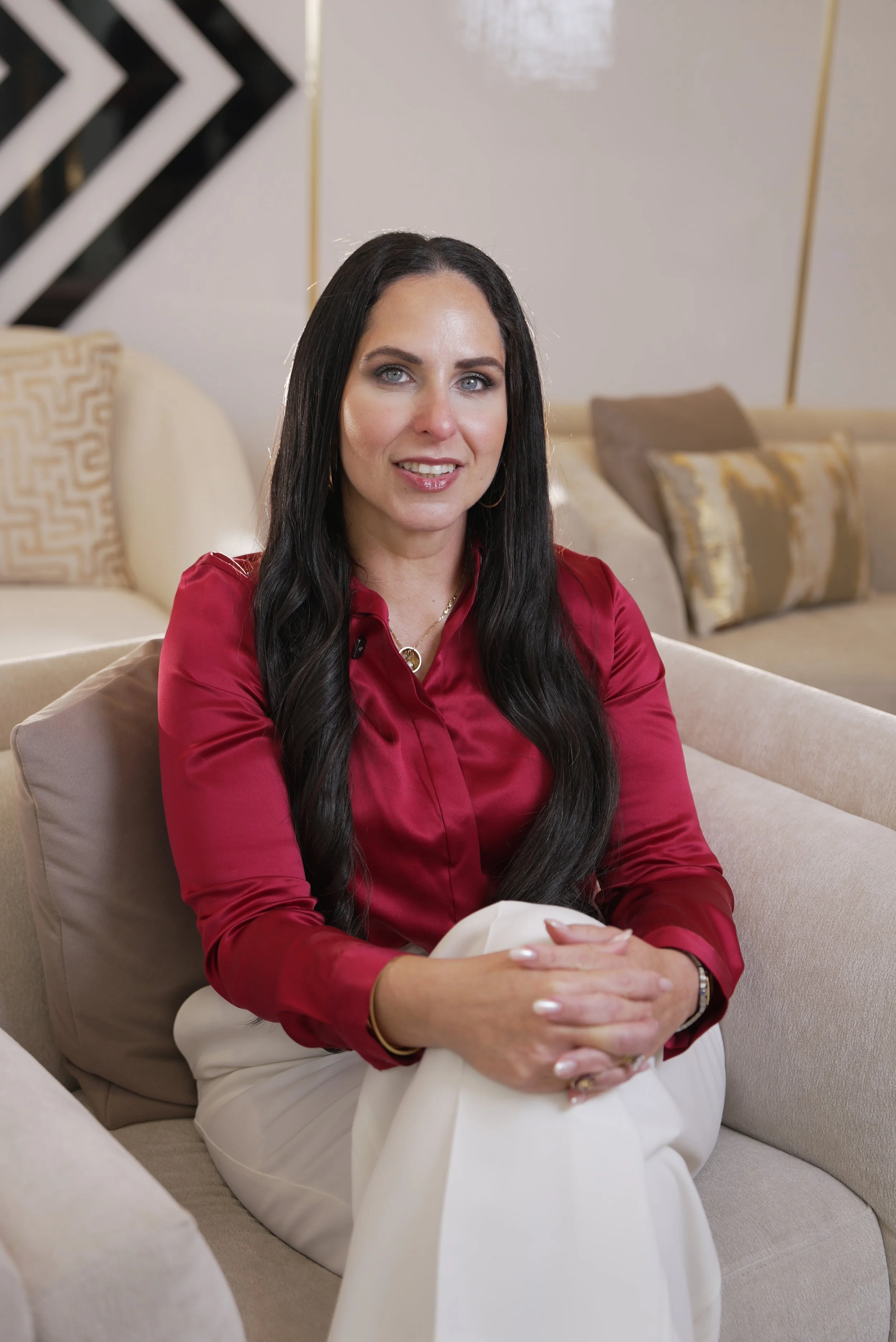 A woman with long dark hair, wearing a red blouse and white pants, sitting on a beige sofa in a well-lit room with decorative pillows and modern wall art.