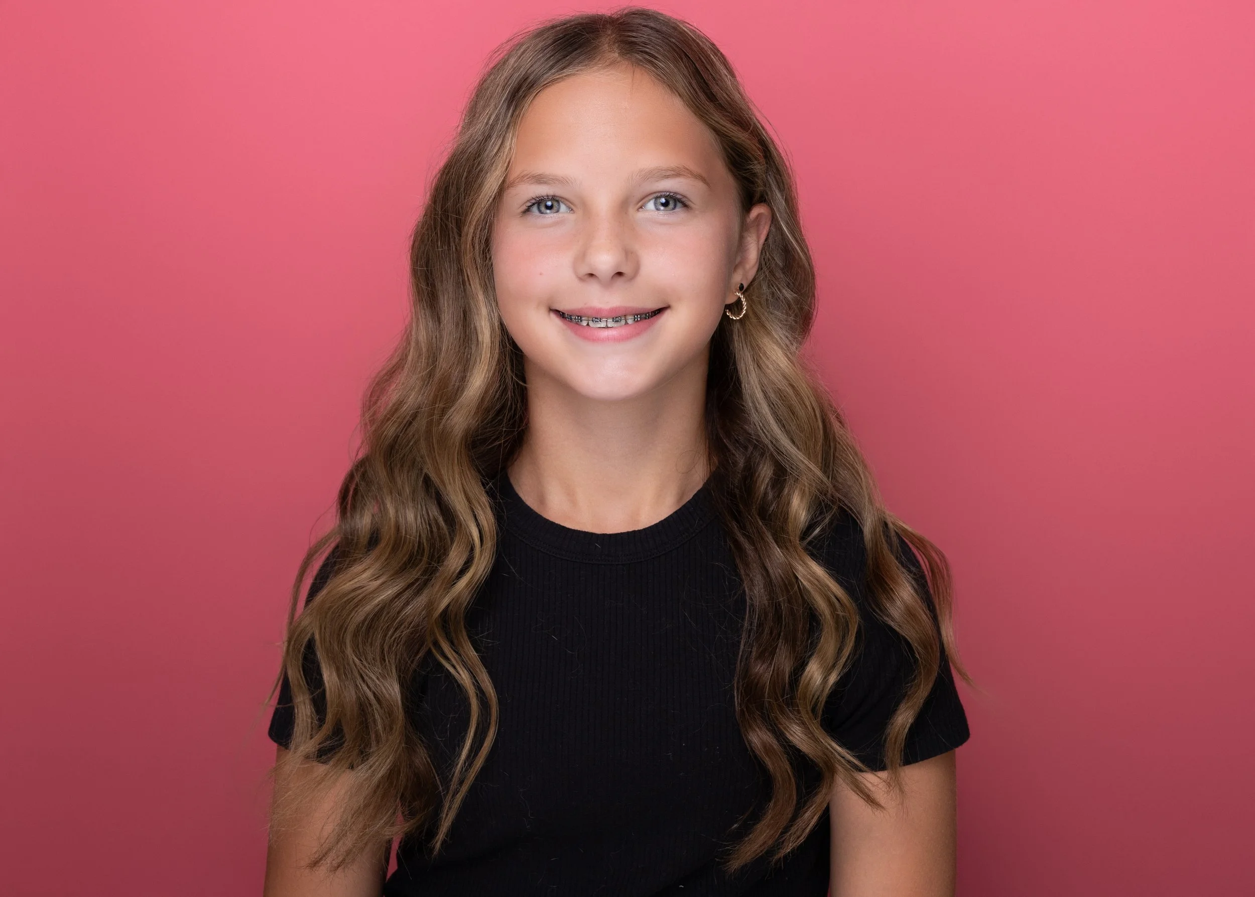 A young girl with long, wavy brown hair, blue eyes, and braces smiling in front of a pink background.