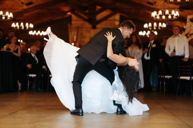 A bride and groom are dancing at their wedding reception, with guests watching in the background.
