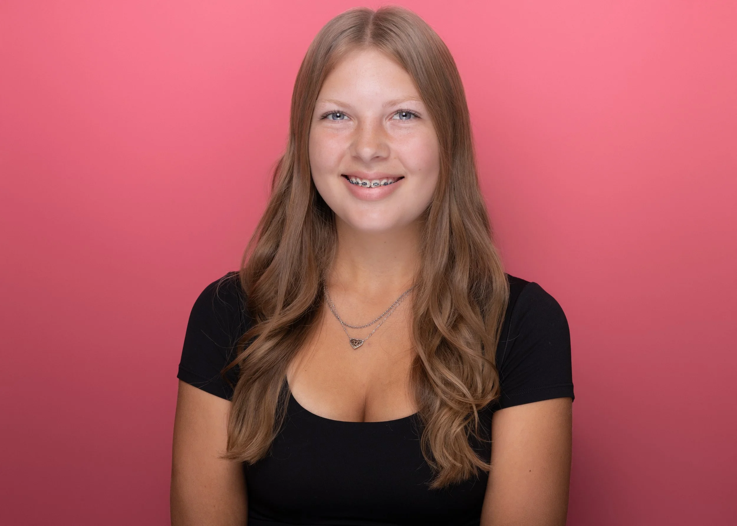 A young woman with long, wavy brown hair and blue eyes, smiling with braces, wearing a black top and two silver necklaces, standing in front of a pink background.