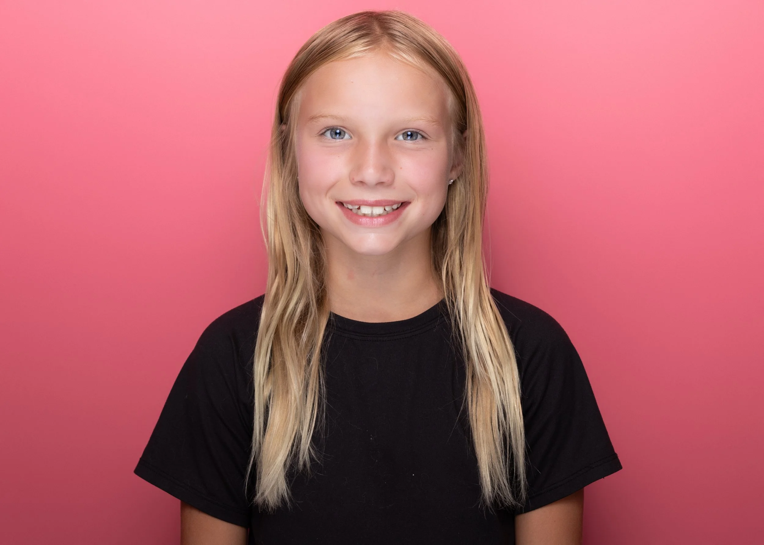 A portrait of a young girl with blonde hair, blue eyes, and a smile, wearing a black shirt, against a pink background.