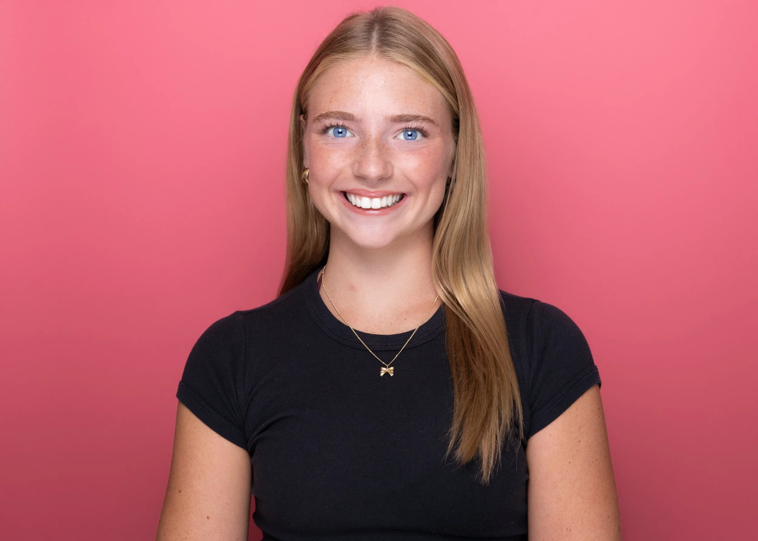 A young woman with long, straight blonde hair and blue eyes smiling in front of a pink background, wearing a black T-shirt and gold jewelry.