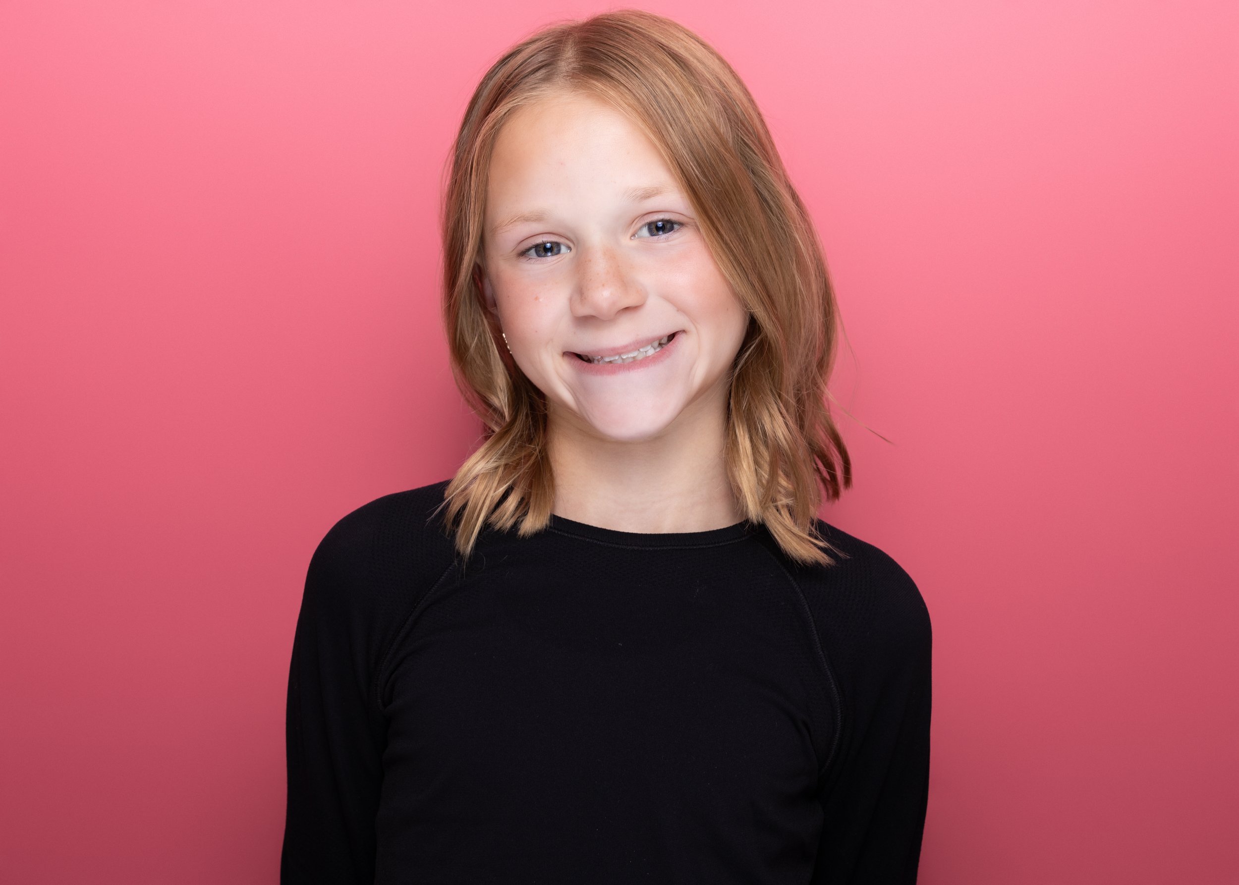 A young girl with red hair, light skin, and blue eyes smiling in front of a pink background, wearing a black long-sleeved shirt.
