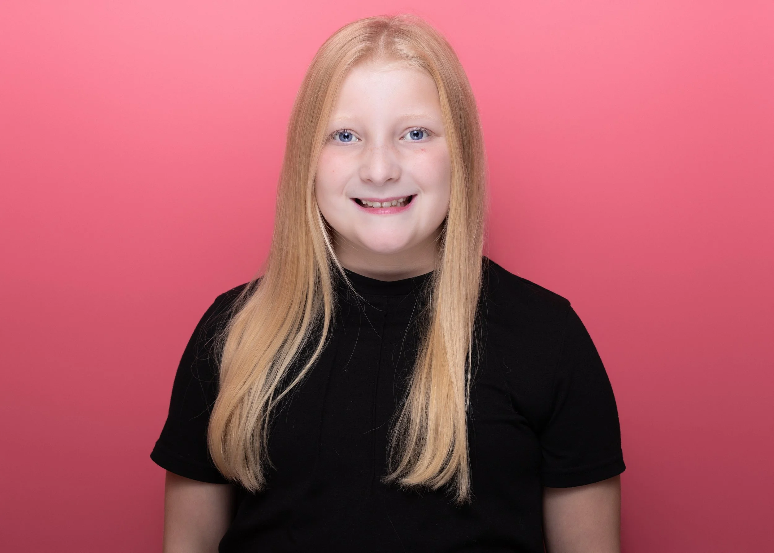 A young girl with long blonde hair, blue eyes, and a small gap in her front teeth, wearing a black shirt, standing against a pink background.