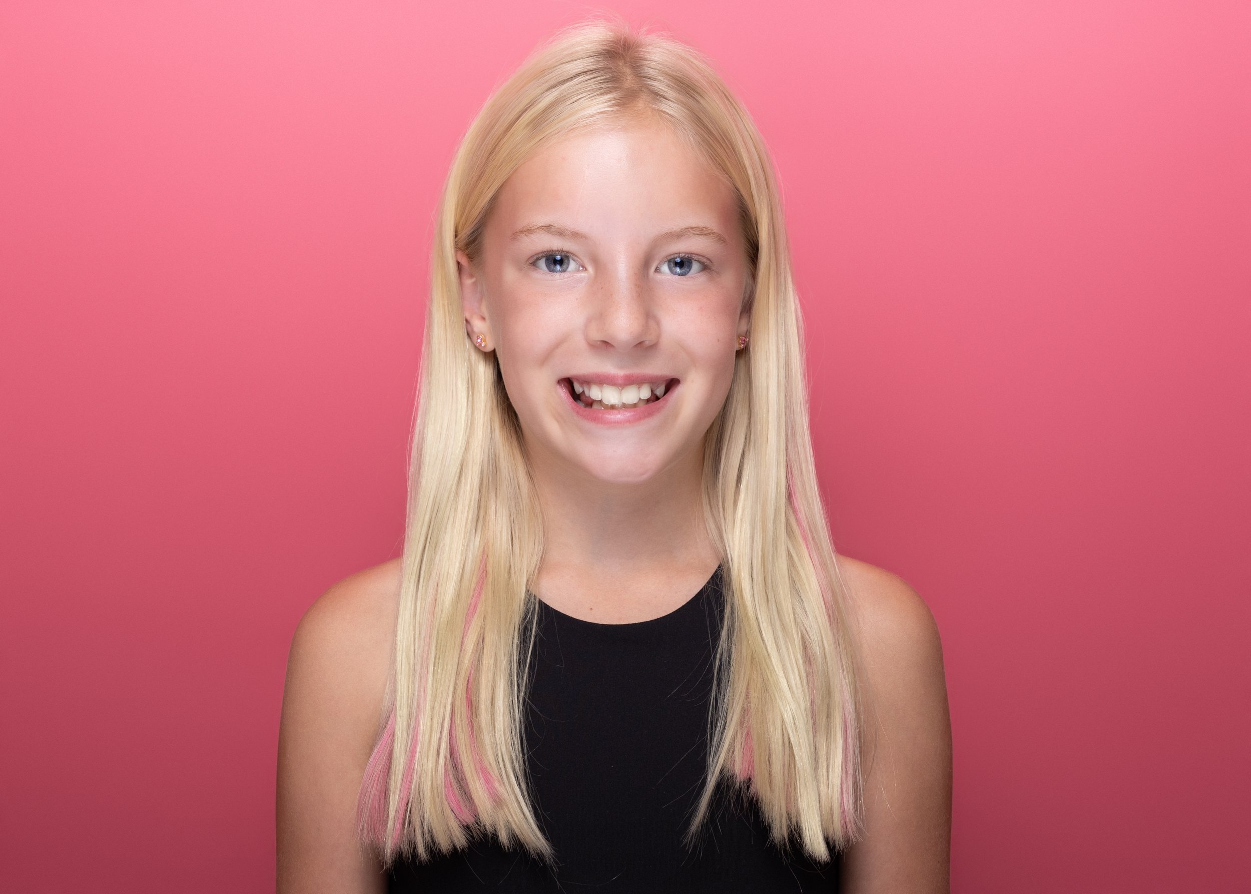 Portrait of a young girl with long blonde hair, wearing a black sleeveless top and pink earrings, smiling against a pink background.