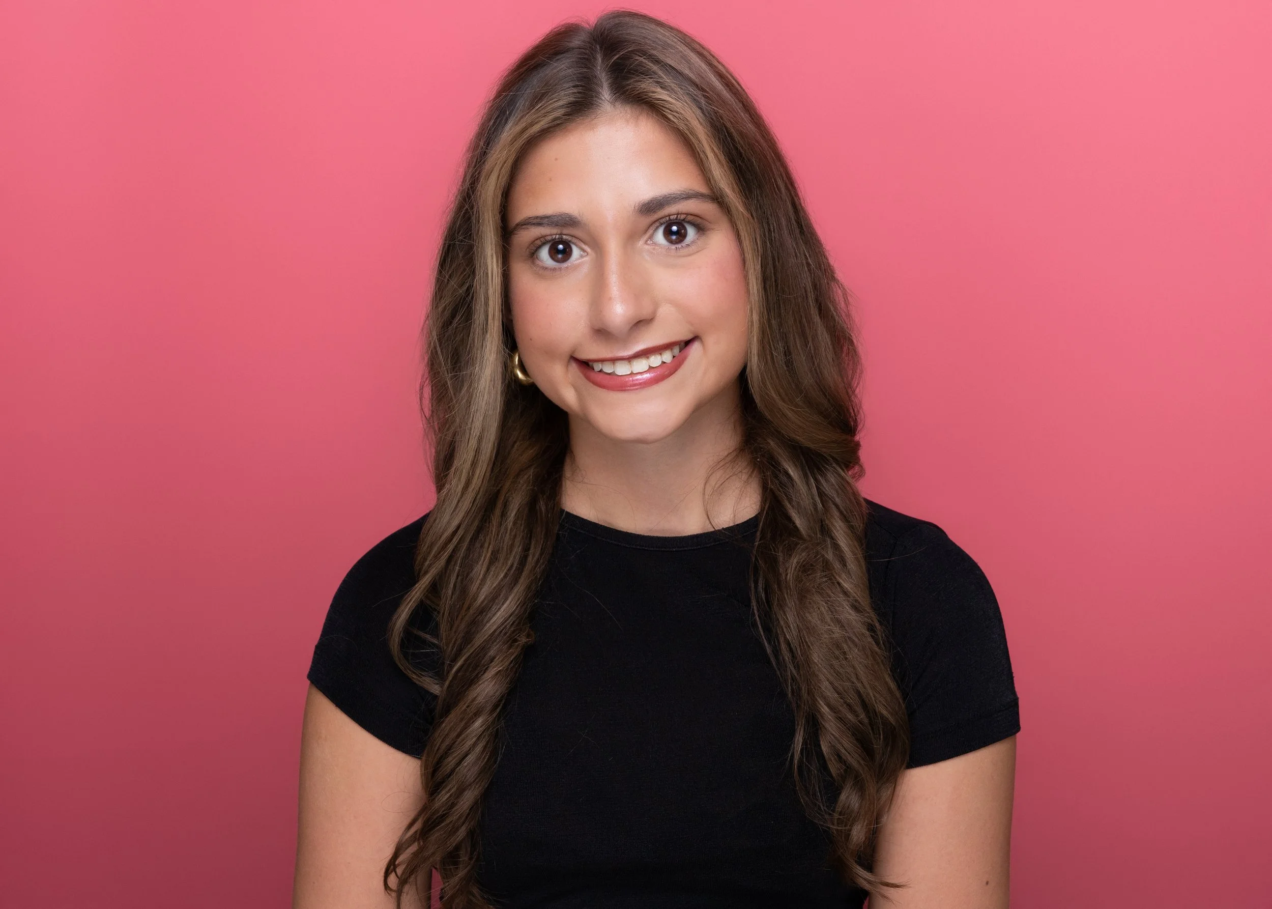 A young woman with long, wavy brown hair and big brown eyes smiling at the camera, wearing a black shirt, against a pink background.
