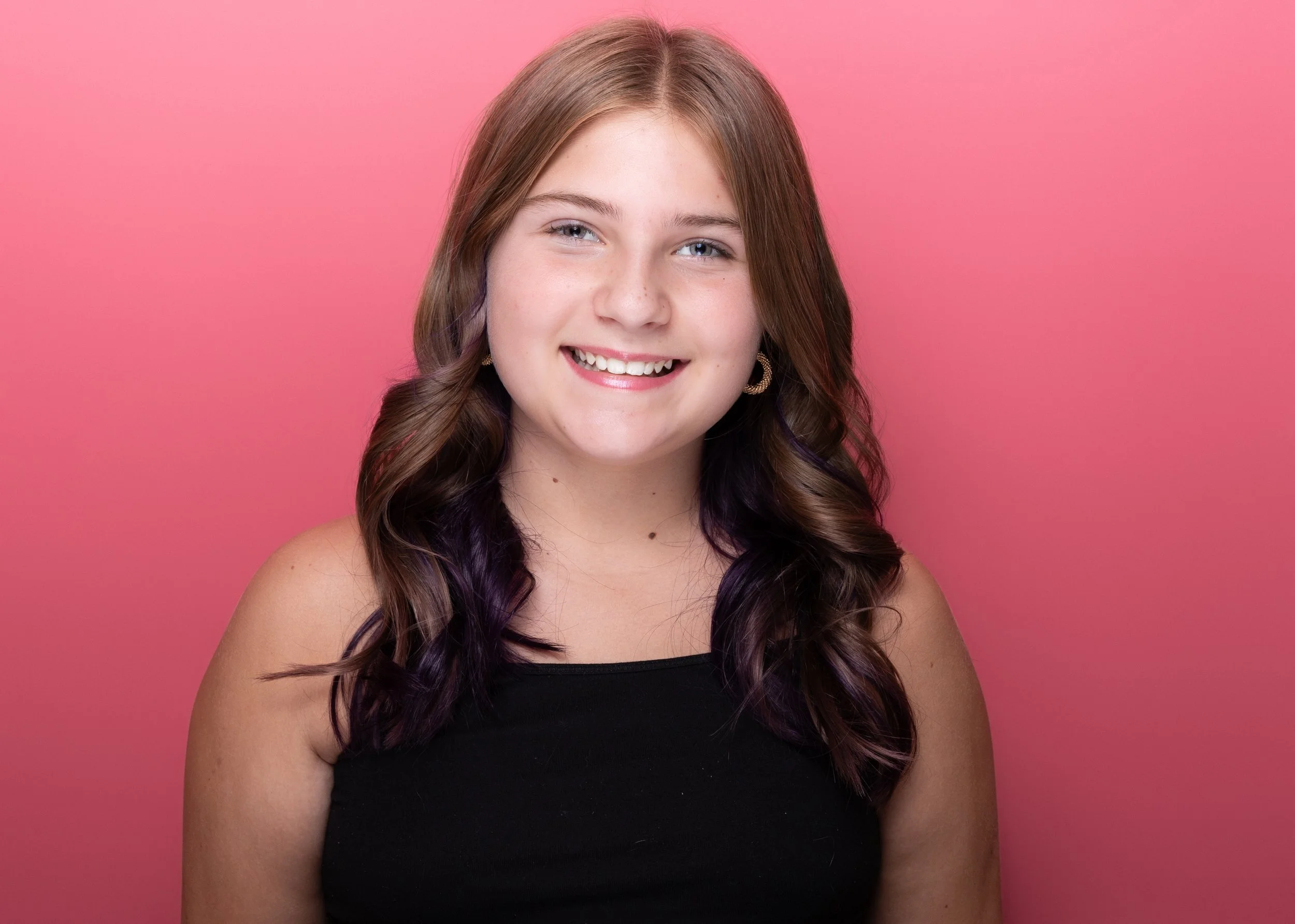 Portrait of a young woman with light skin and long brown and purple hair, smiling, against a pink background.
