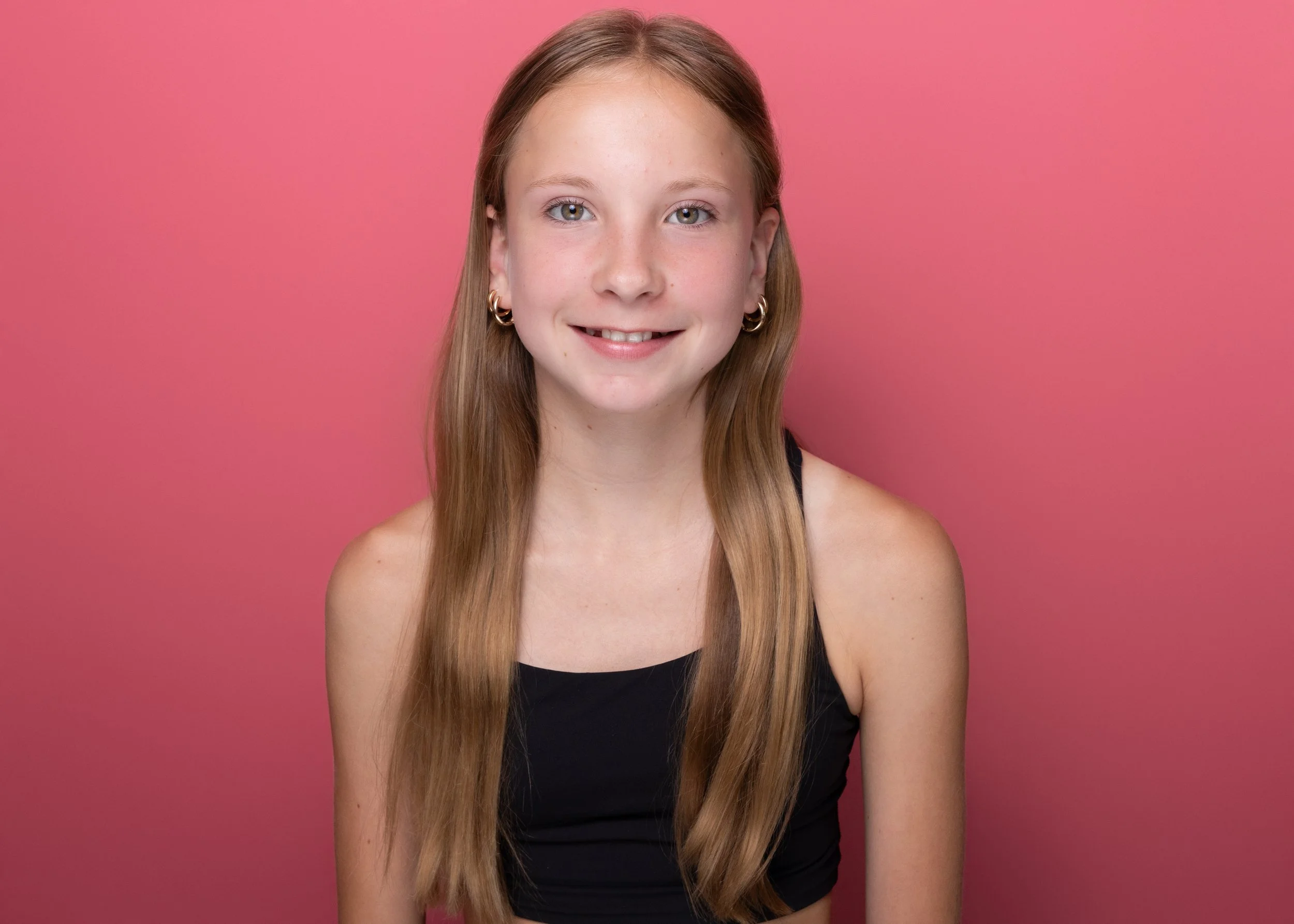 Portrait of a young girl with long, straight, reddish-brown hair, wearing hoop earrings and a black top, smiling in front of a pink background.