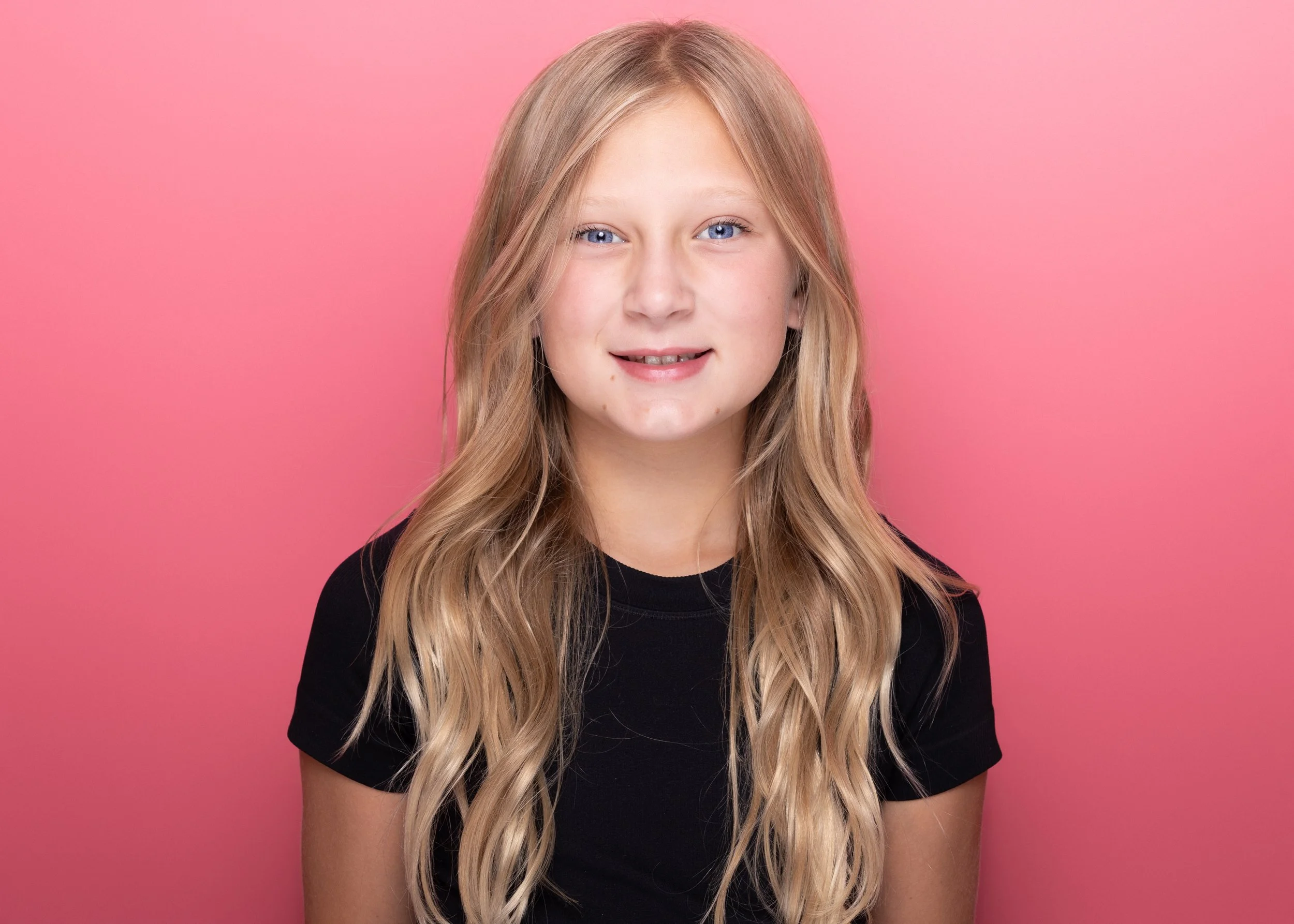 A young girl with long, wavy blonde hair and blue eyes, wearing a black shirt, standing against a pink background.