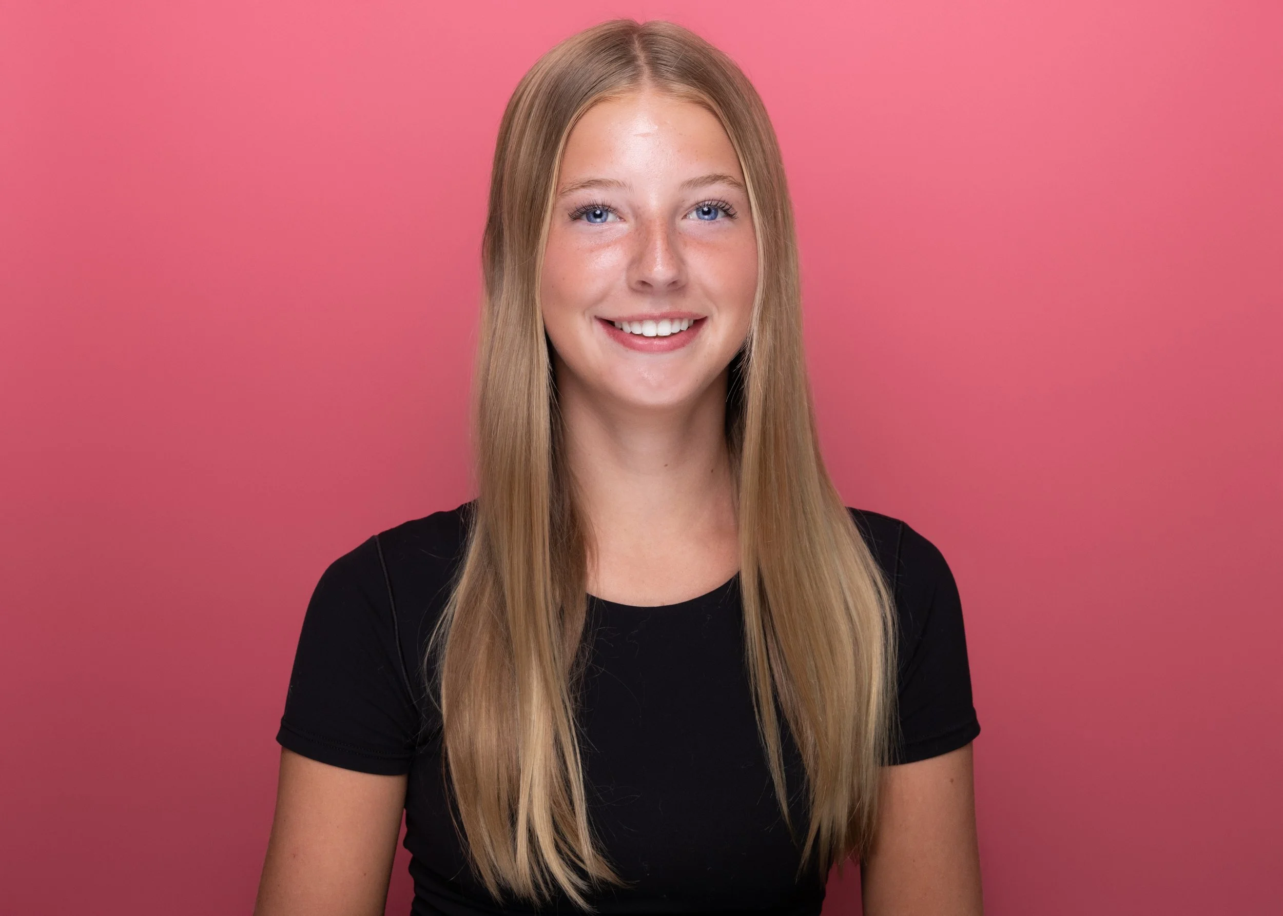 A young woman with long blonde hair, blue eyes, and freckles smiling in front of a pink background, wearing a black top.