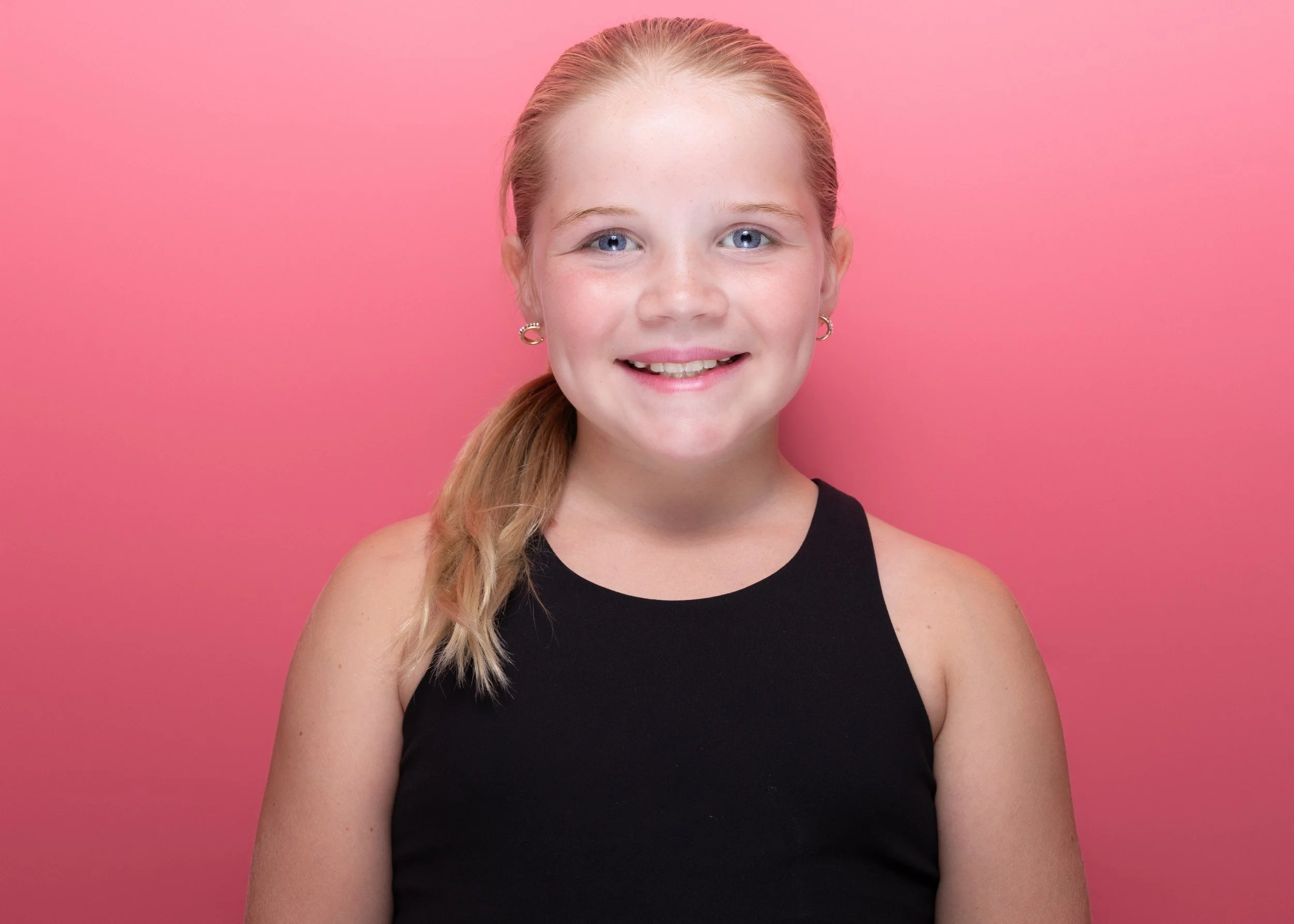 A young girl with blue eyes, light red hair tied back, wearing a black sleeveless top with earrings, smiling in front of a solid pink background.