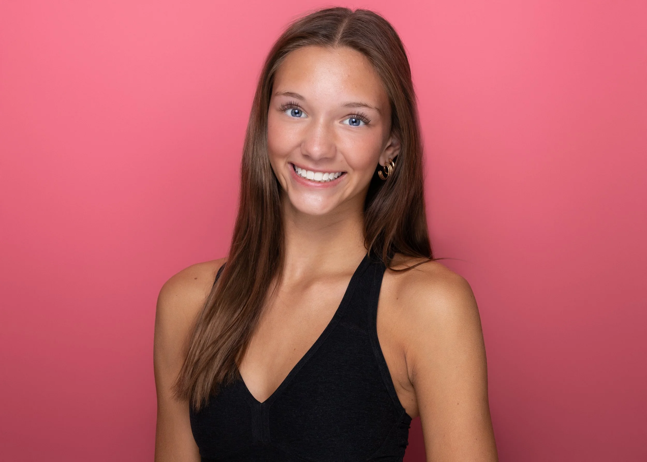 A young woman with long brown hair, blue eyes, and a big smile, wearing golden hoop earrings and a black sleeveless top, standing against a pink background.