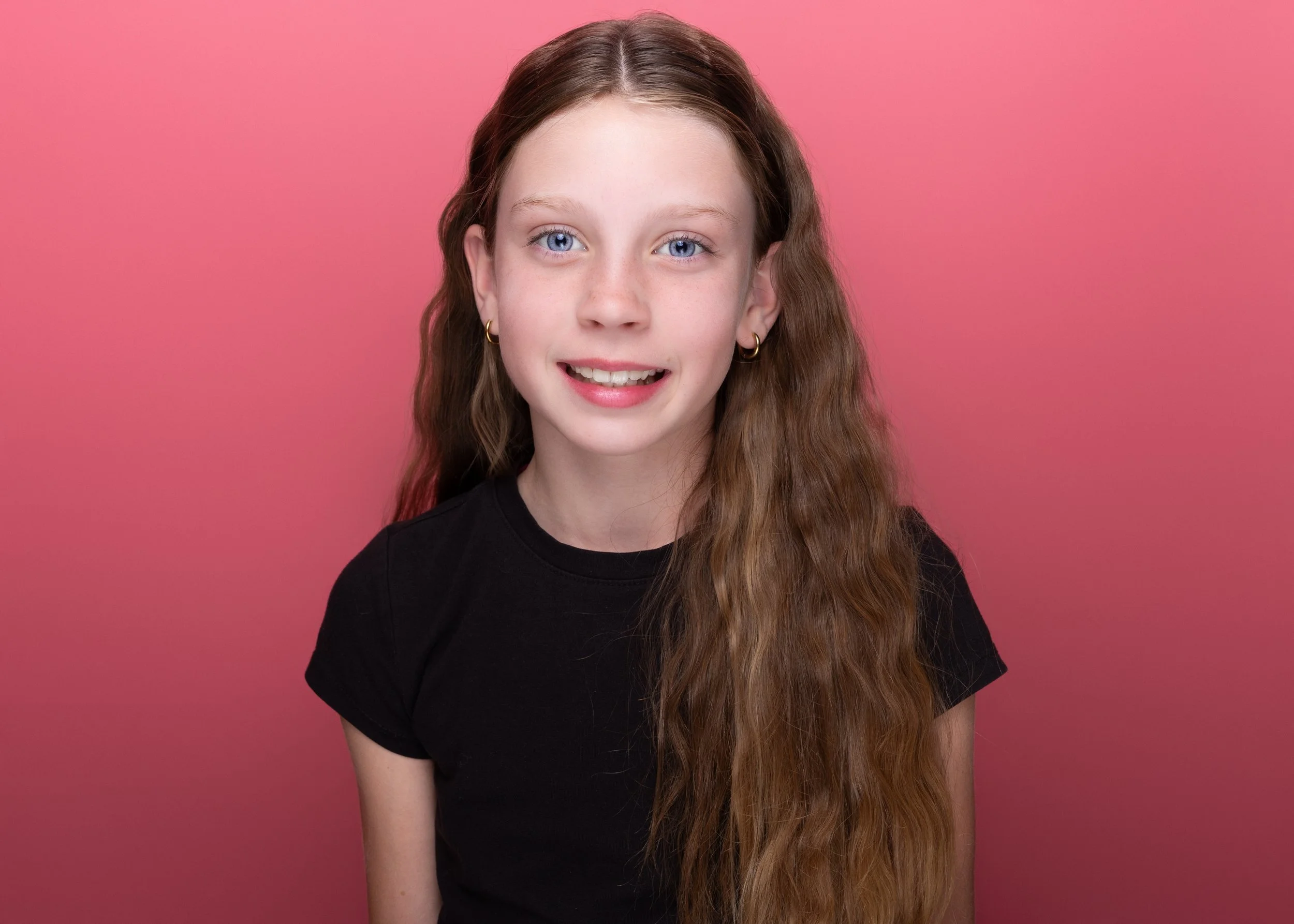 Portrait of a young girl with long, wavy light brown hair and blue eyes, wearing a black t-shirt and small gold hoop earrings, standing against a pink background.