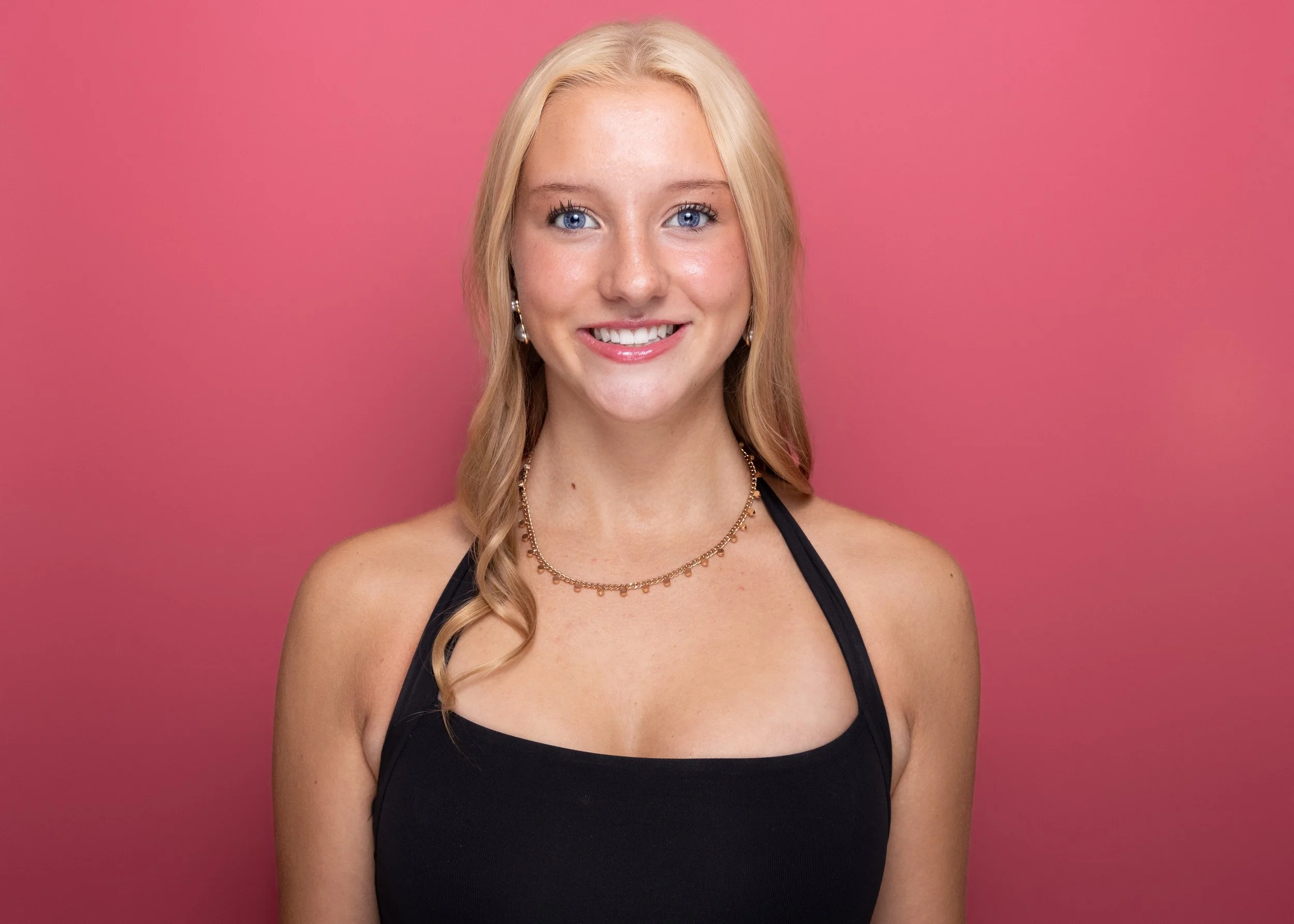 A smiling woman with blonde hair, blue eyes, wearing earrings, a gold necklace, and a black tank top, standing against a pink background.