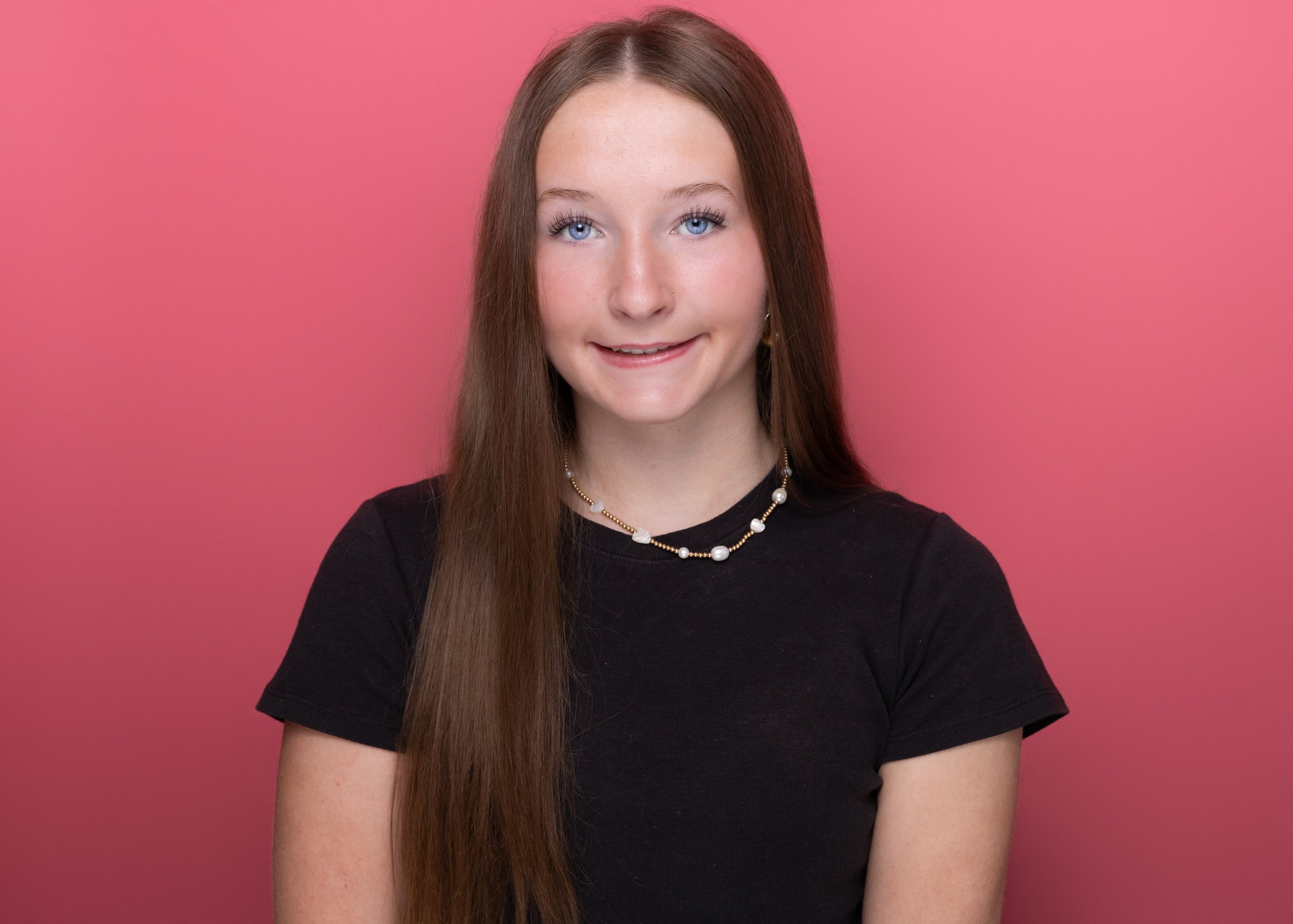 Portrait of a young woman with long brown hair, blue eyes, smiling, wearing a black shirt and a pearl jewelry necklace, against a pink background.