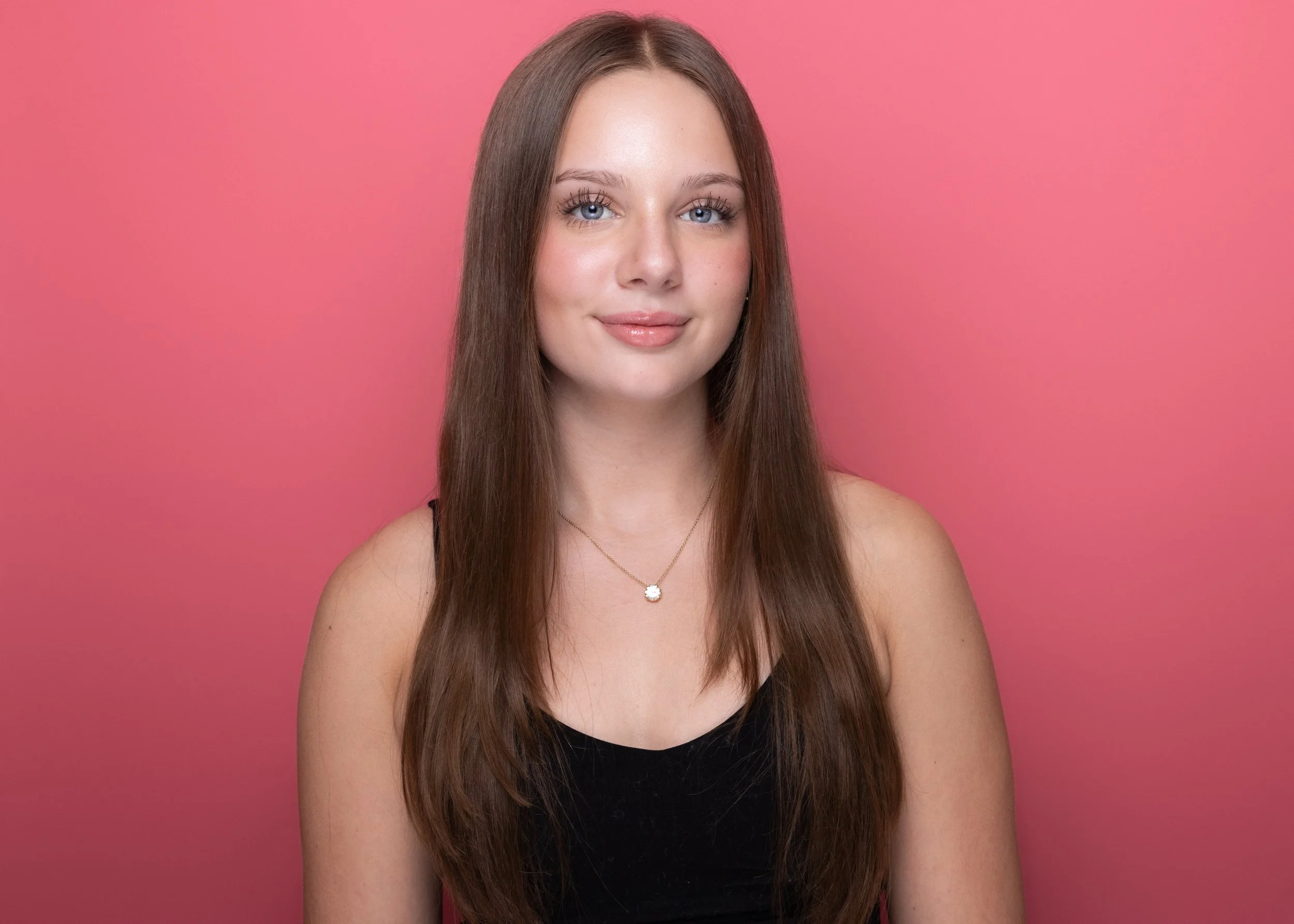 Portrait of a young woman with long brown hair and blue eyes, wearing a black top and a necklace with a white pendant, set against a pink background.