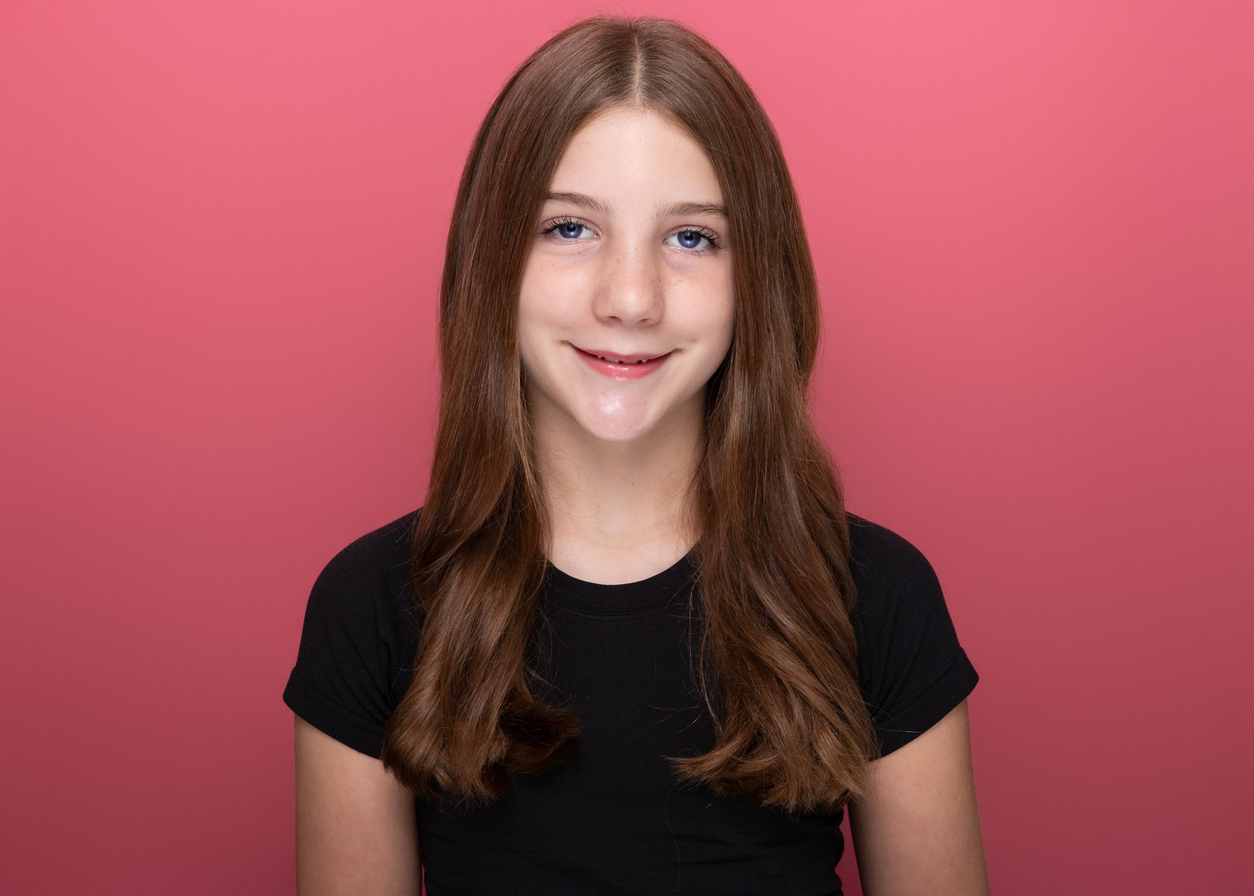 A young woman with long, wavy light brown hair and blue eyes smiling in front of a pink background, wearing a black t-shirt.