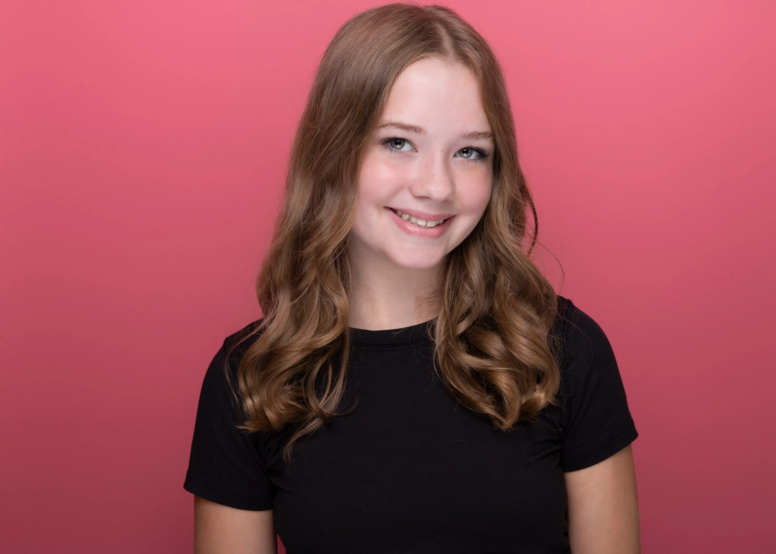 A young woman with light skin, long wavy auburn hair, blue eyes, and freckles wearing a black shirt, smiling in front of a pink background.