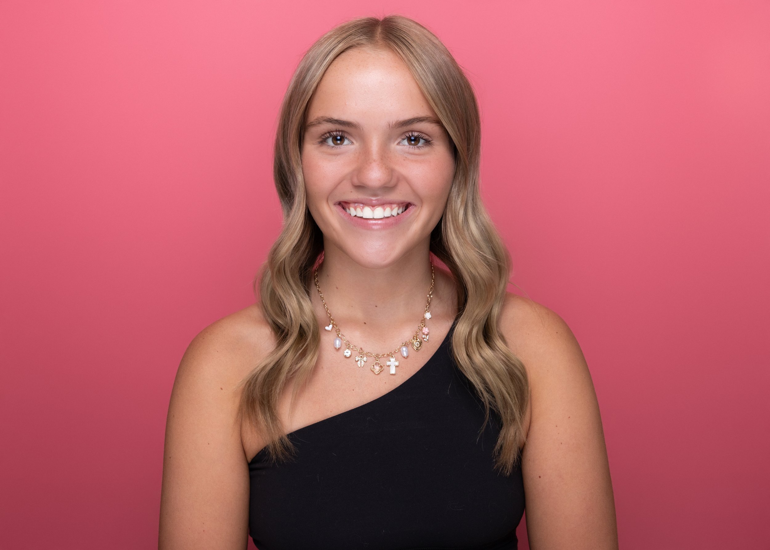 A young woman with long, wavy blonde hair and light skin, smiling, wearing a black one-shoulder top, colorful jewelry including necklaces with a cross and angel charm, earrings, and a necklace with pearls and charms, standing against a pink backgroun