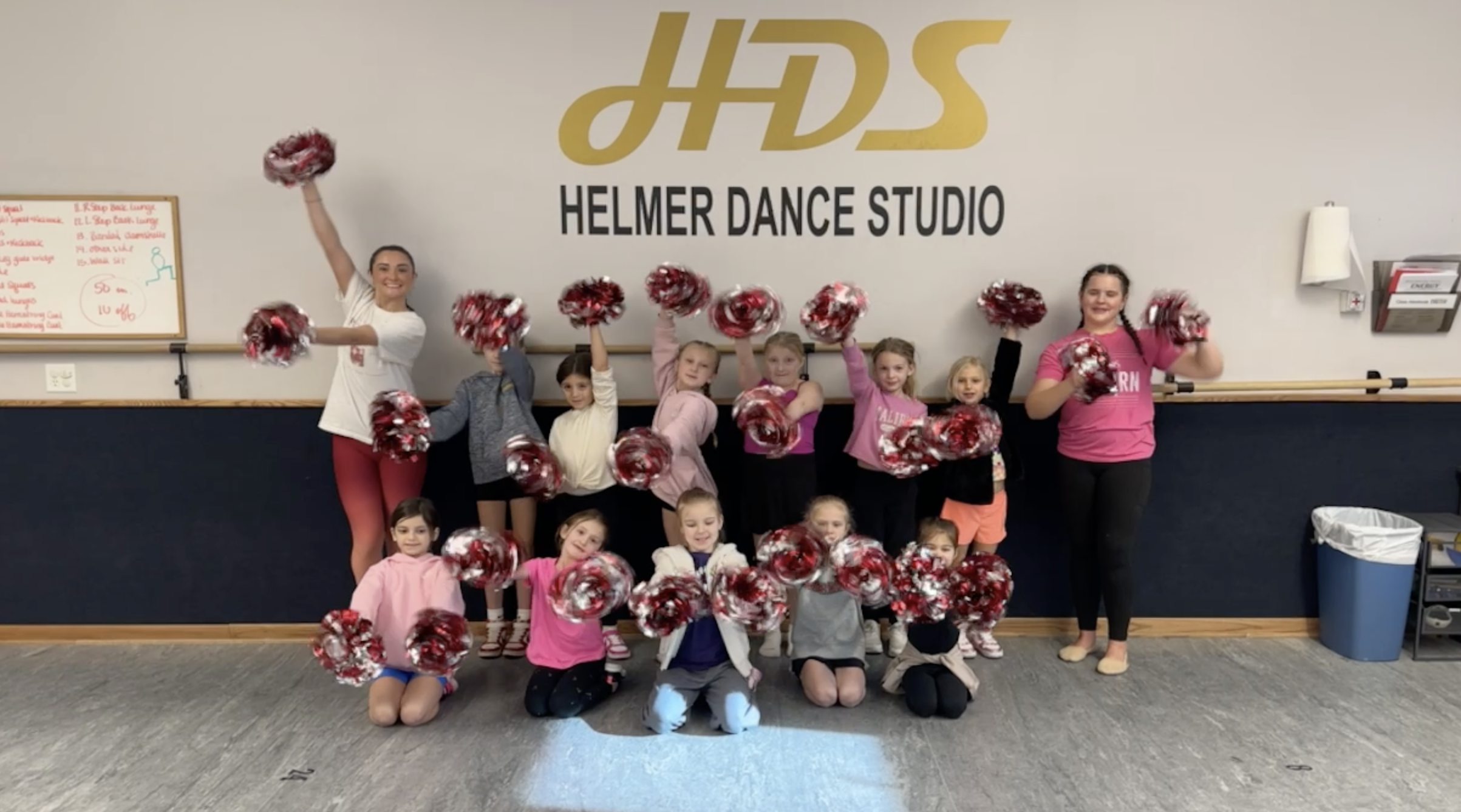 Group of children and two instructors at Helmer Dance Studio, holding red and white pom-poms, posing for a photo in a dance studio.
