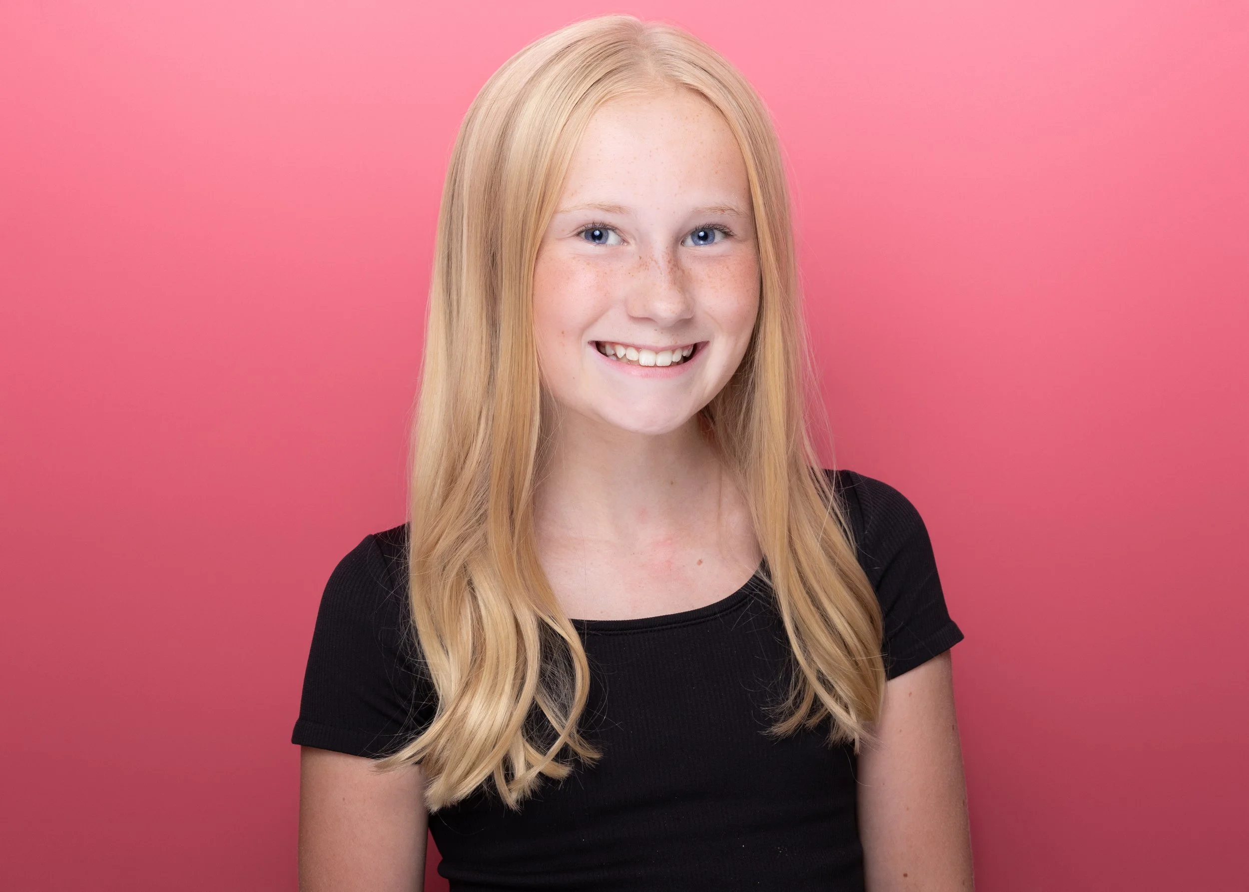 A young woman with long blonde hair, blue eyes, and freckles, smiling, against a pink background, wearing a black shirt.