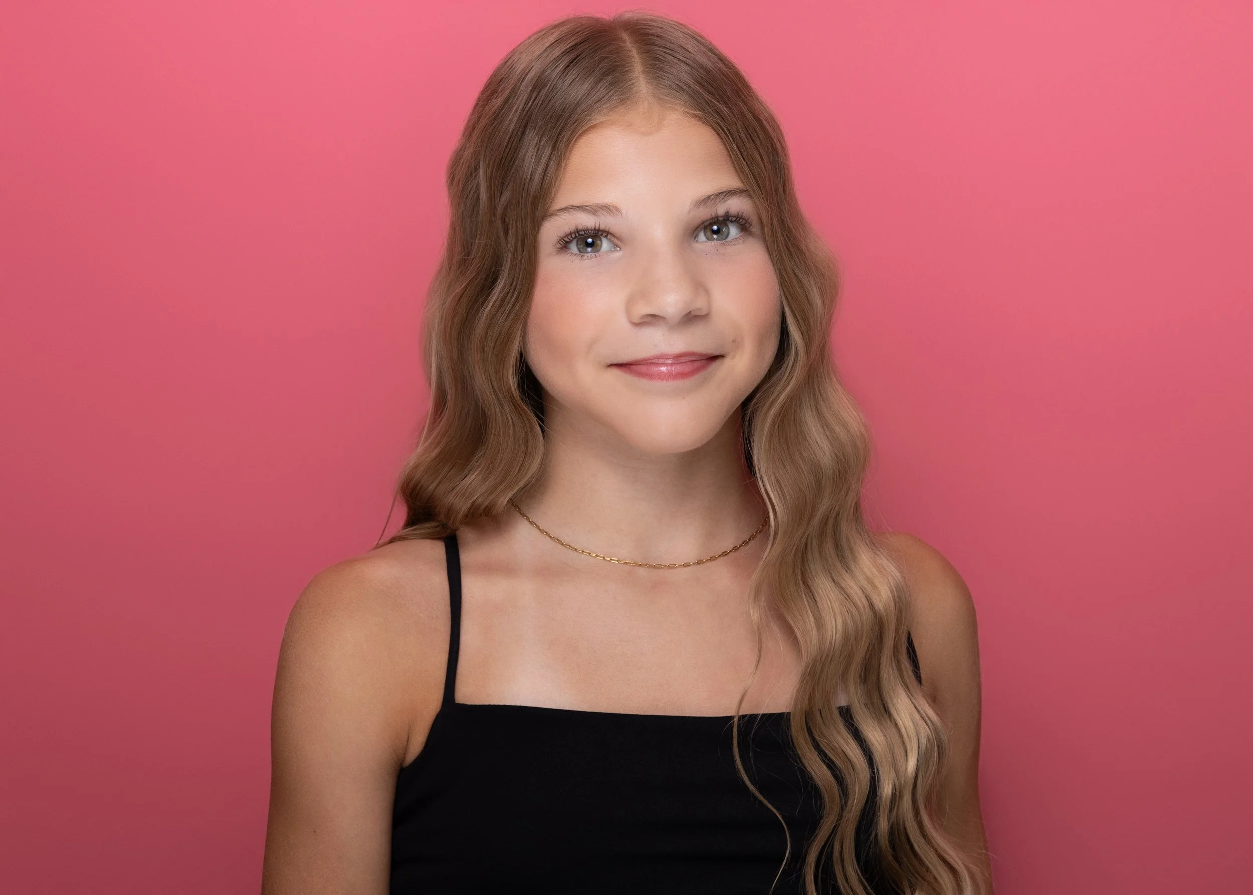 A young woman with long, wavy blonde hair and light skin, smiling slightly, wearing a black spaghetti strap top and a delicate gold necklace, against a solid pink background.